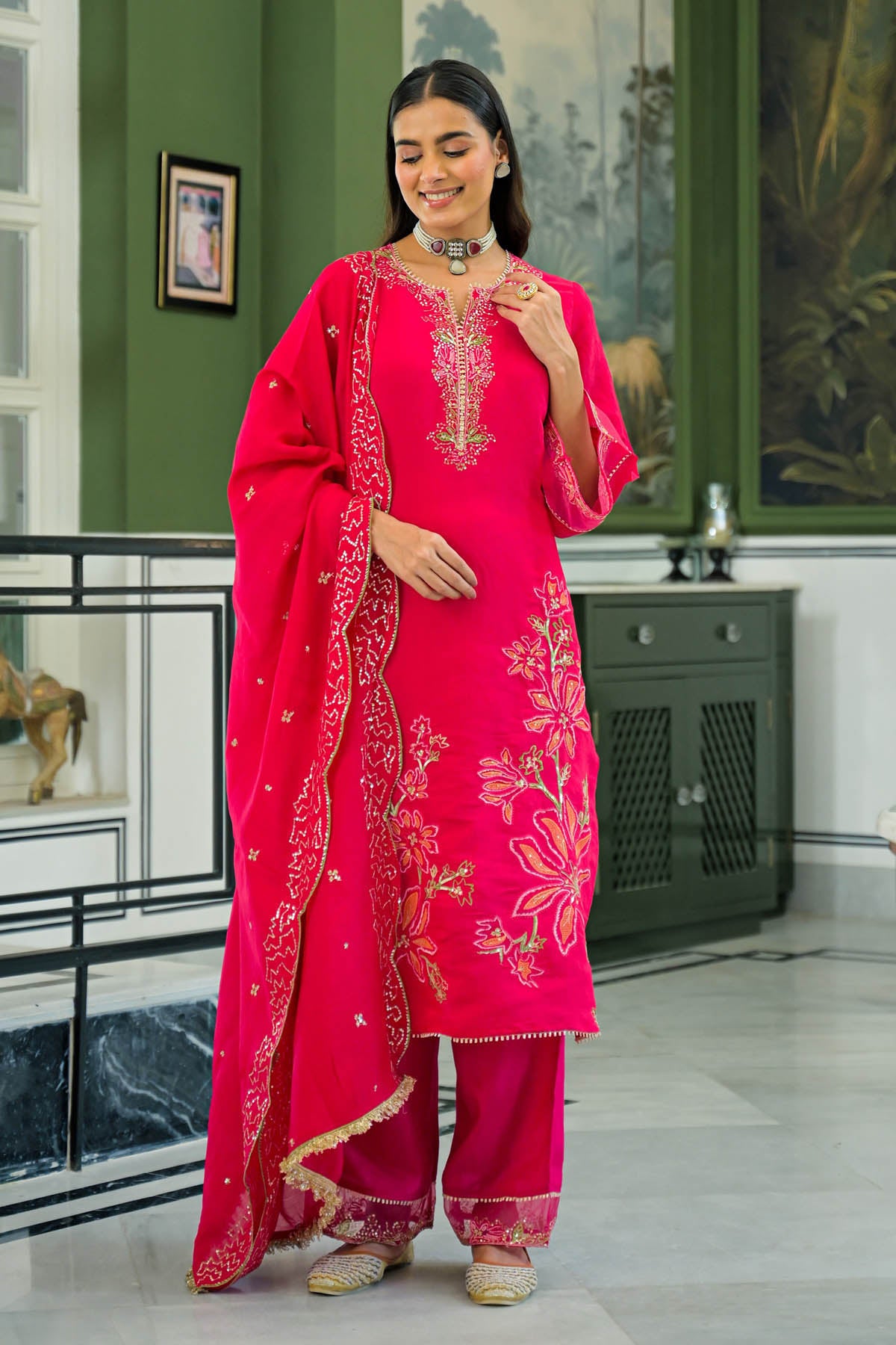Woman in a bright pink traditional outfit with intricate designs in a room with green walls and decorative items.