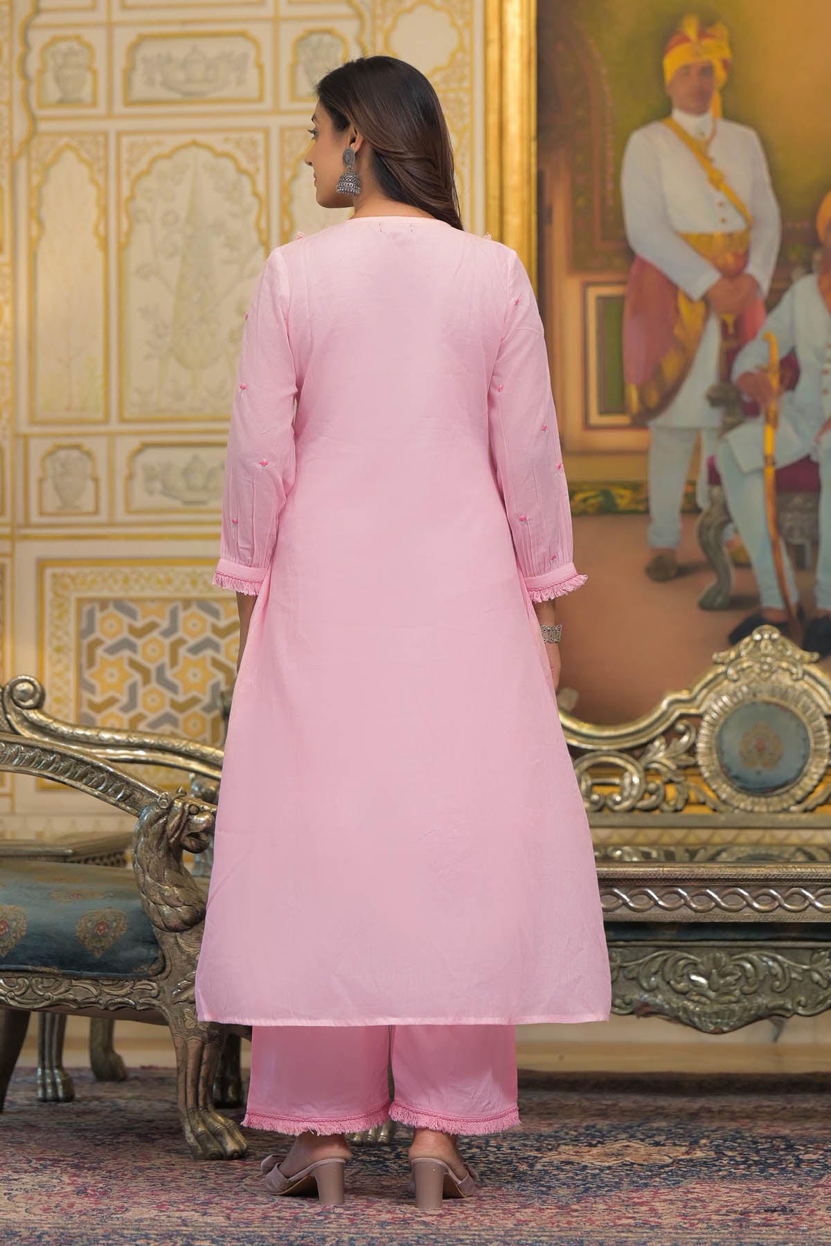 Woman in a pink traditional outfit standing in an ornate room with decorative walls and furniture.