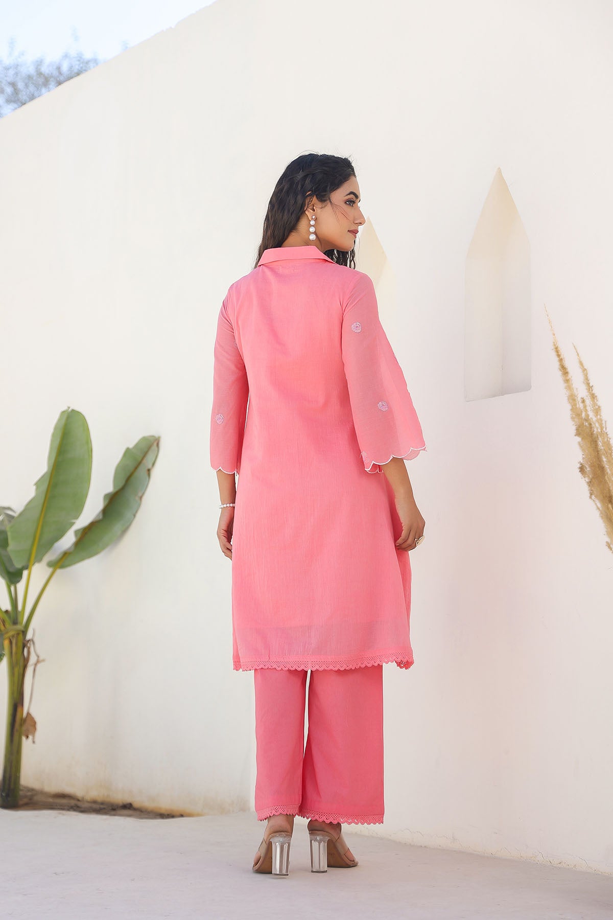 Woman wearing a pink outfit standing against a white wall with plants around.