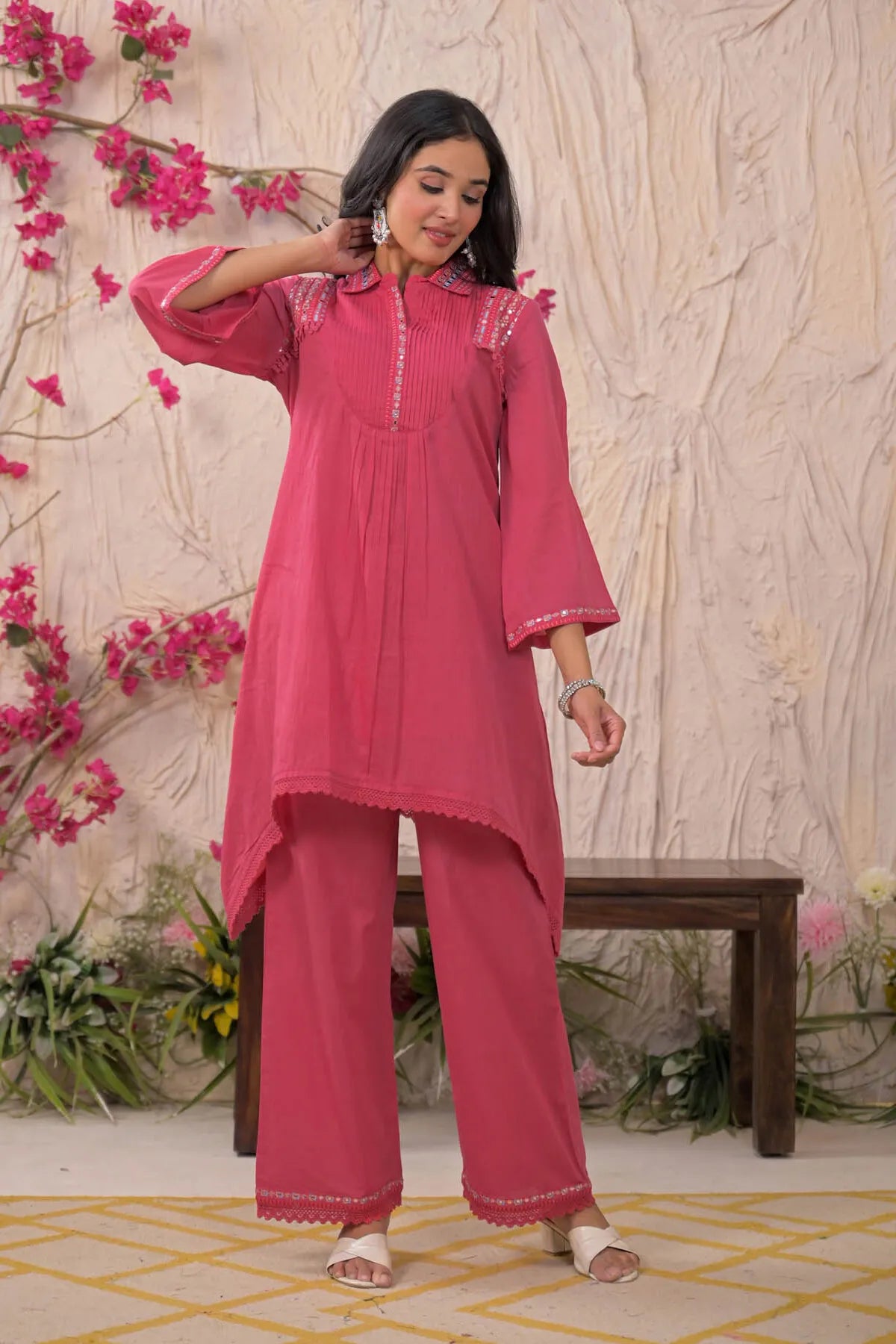 Woman wearing a pink traditional outfit with floral decorations against a decorative wall.