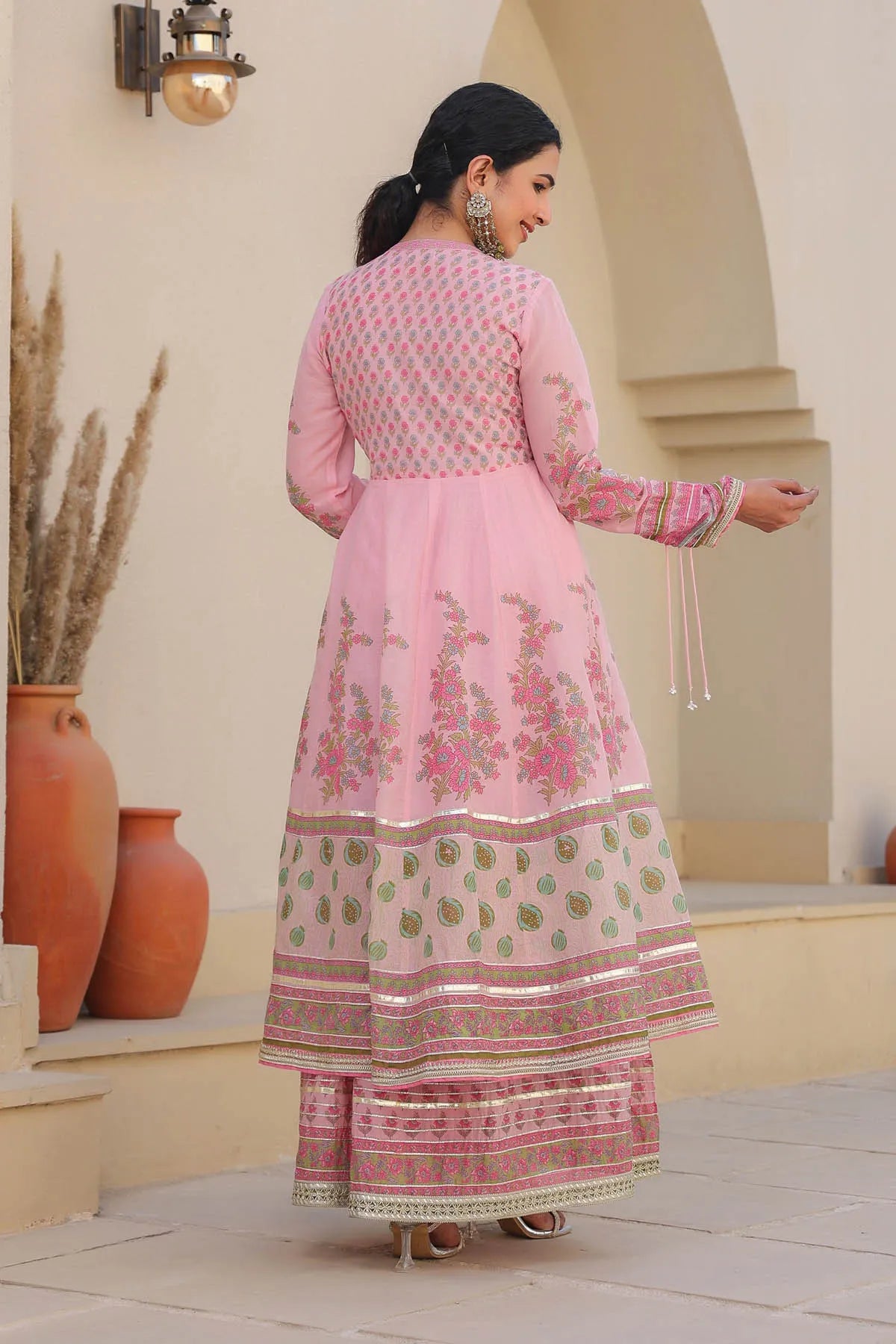 Woman in a pink traditional outfit standing outdoors with decorative vases and archway in the background.