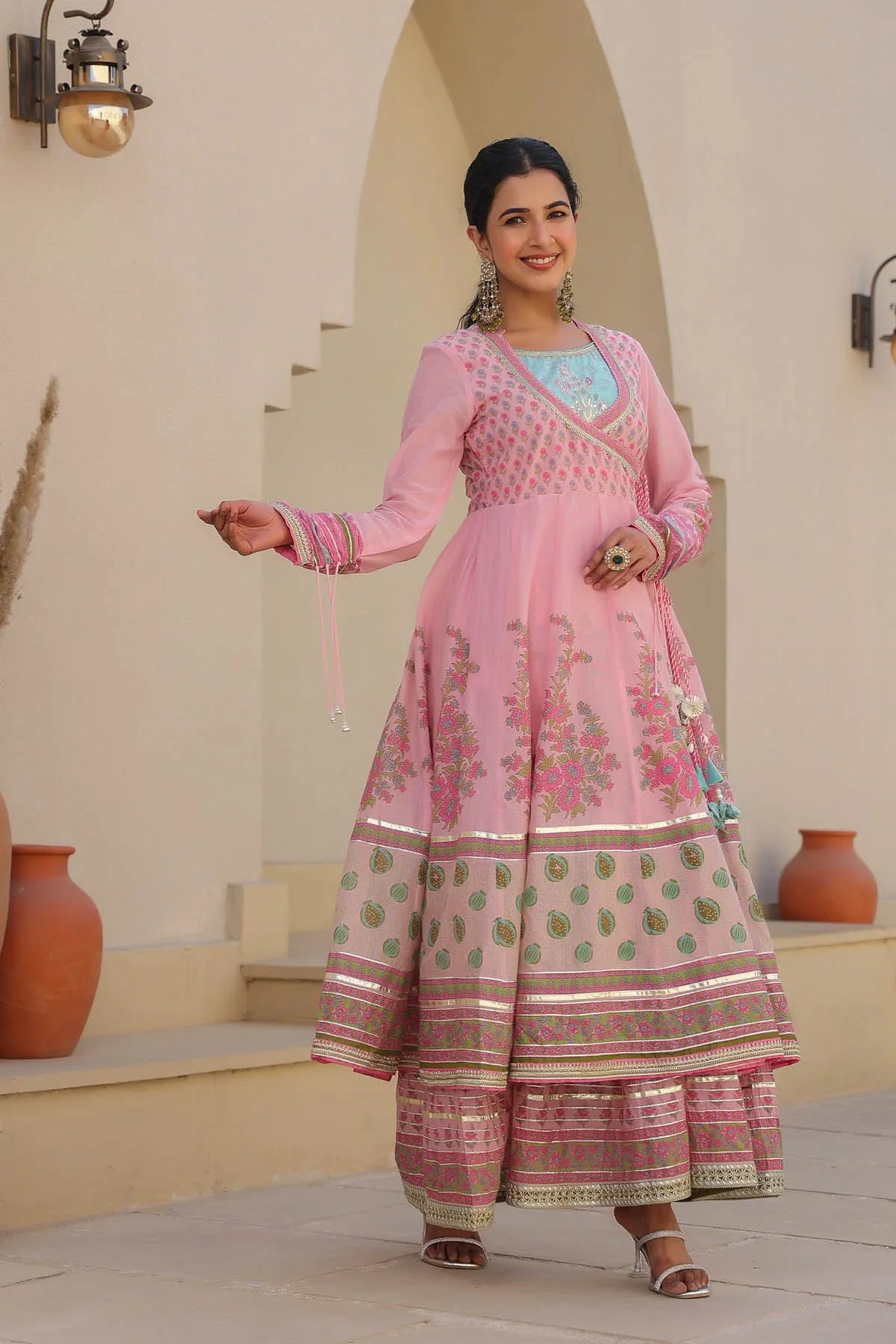 Woman in a pink traditional outfit standing in an outdoor setting with beige walls and terracotta pots.