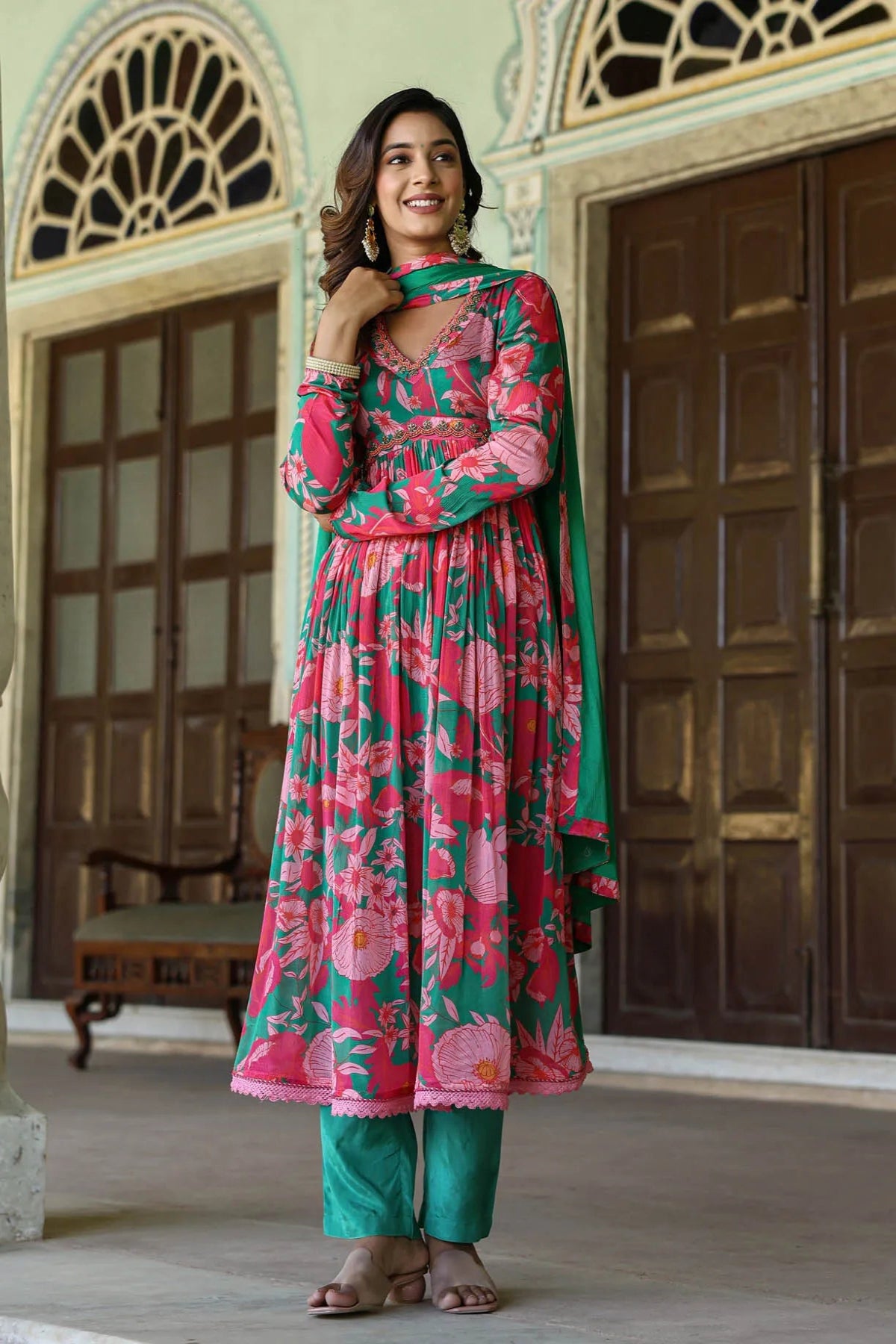 Woman in a pink and green floral outfit standing in an ornate indoor setting.