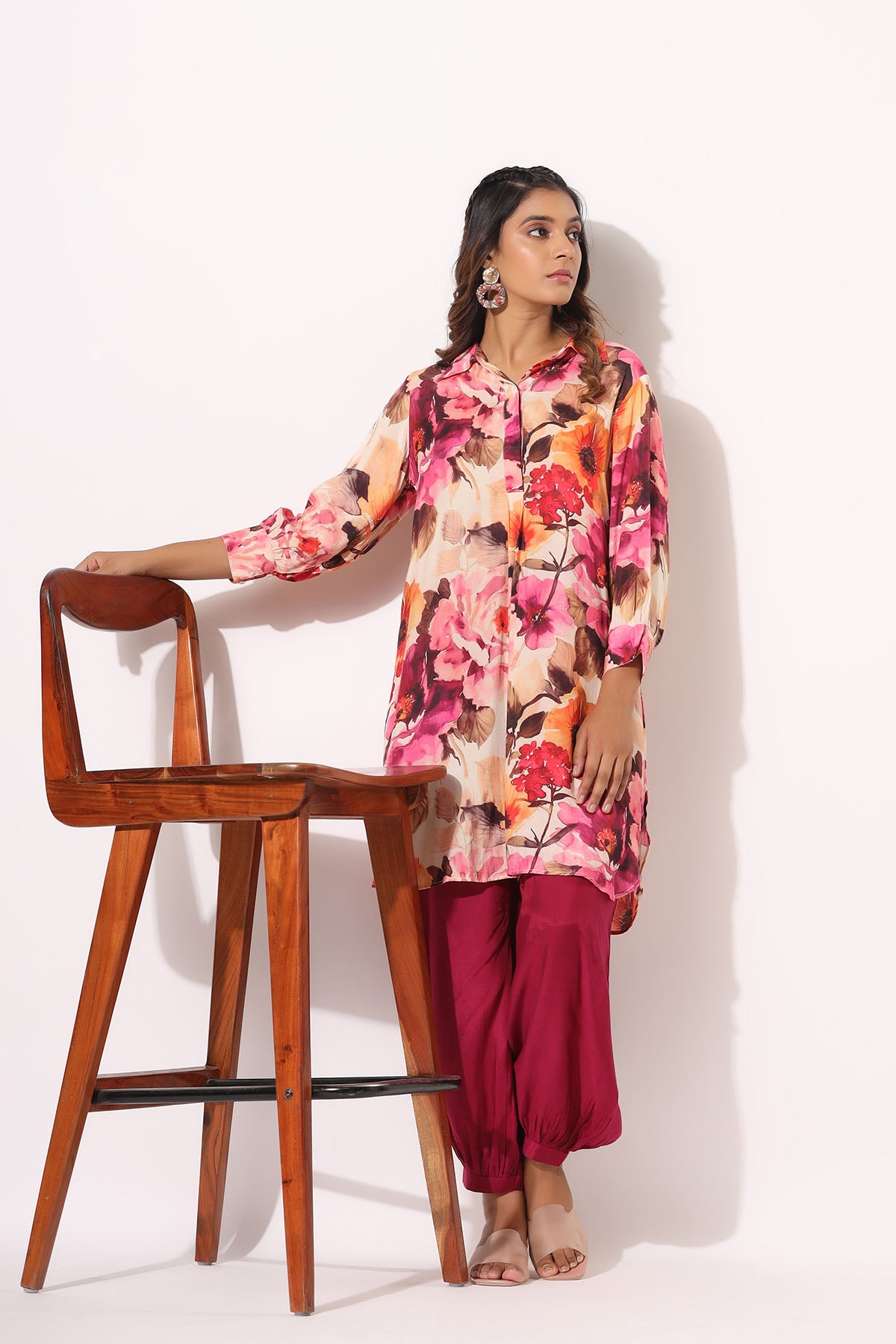 Woman wearing a floral top and pink pants leaning against a wooden stool on a white background