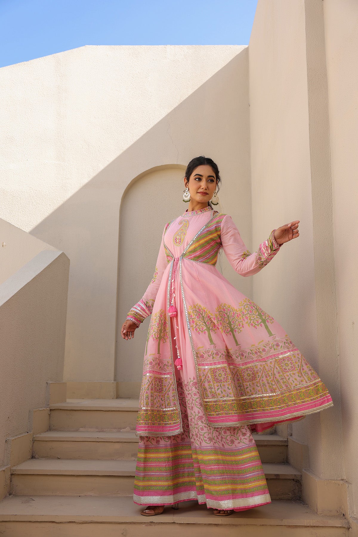 Woman in a pink traditional outfit standing on steps with a beige wall and blue sky in the background