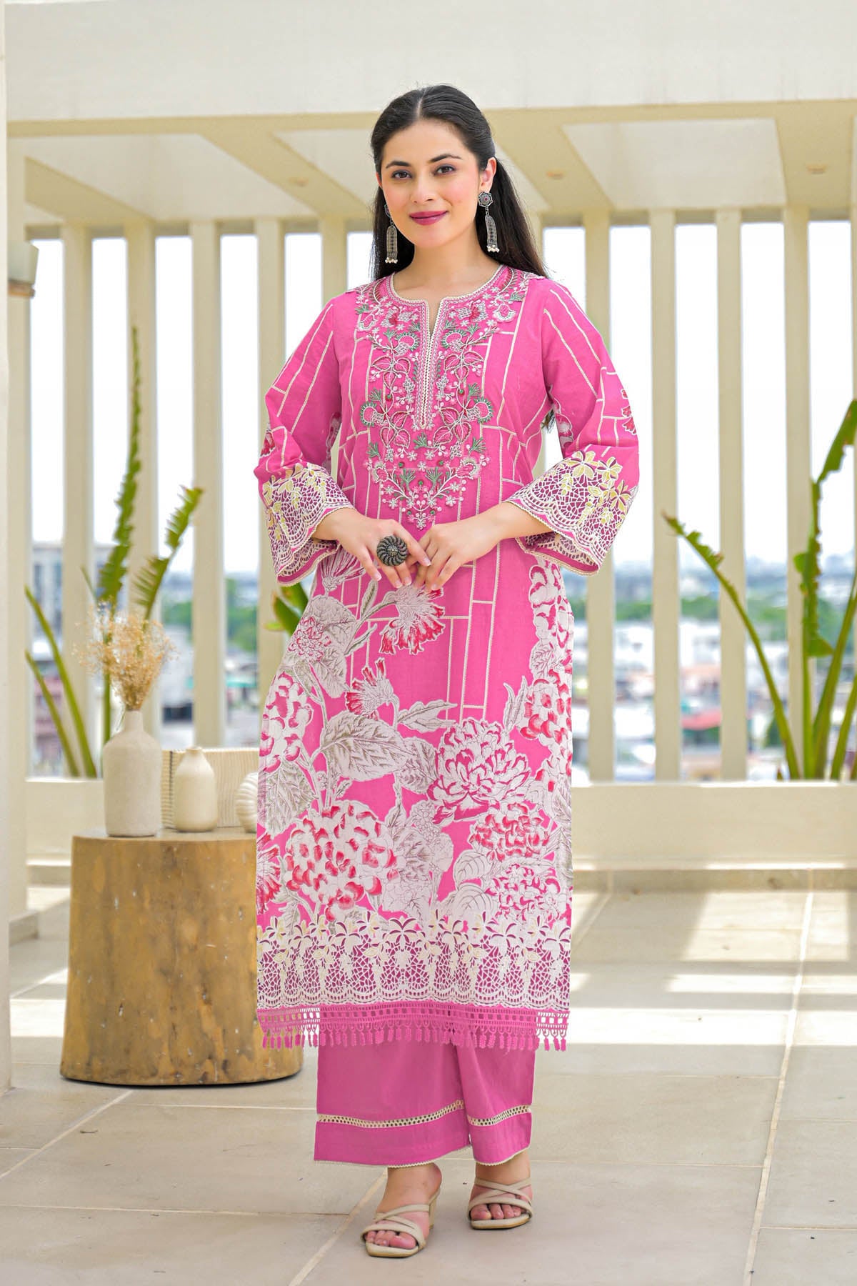 Woman wearing a pink embroidered outfit on a balcony with plants and a city view.