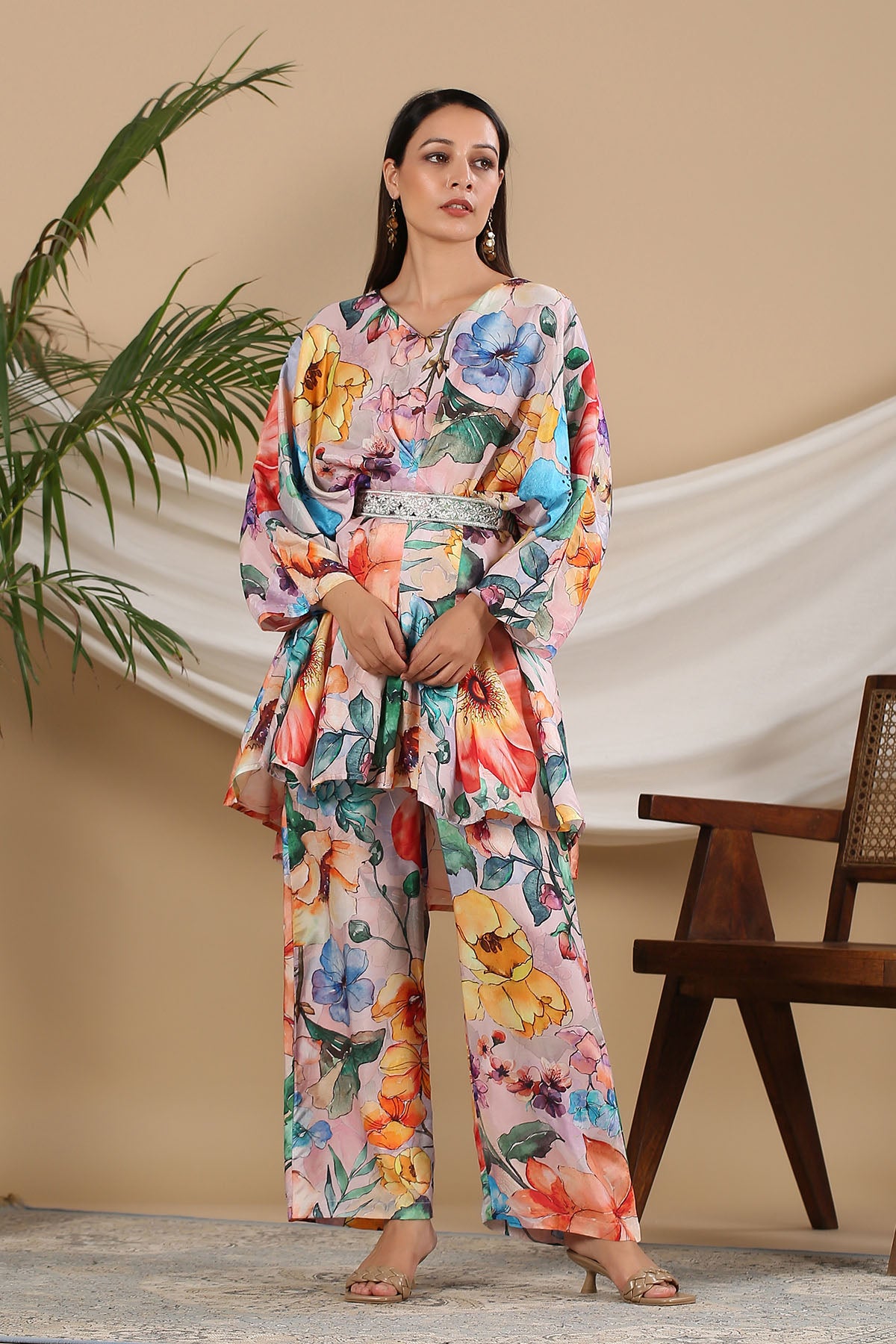 Woman wearing a colorful floral outfit standing in a room with a plant and wooden chair.