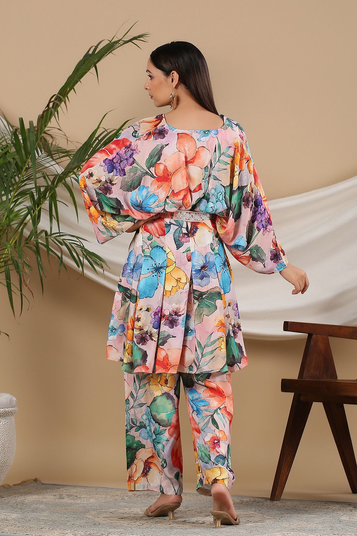 Woman wearing a colorful floral outfit standing in a room with a plant and wooden bench.