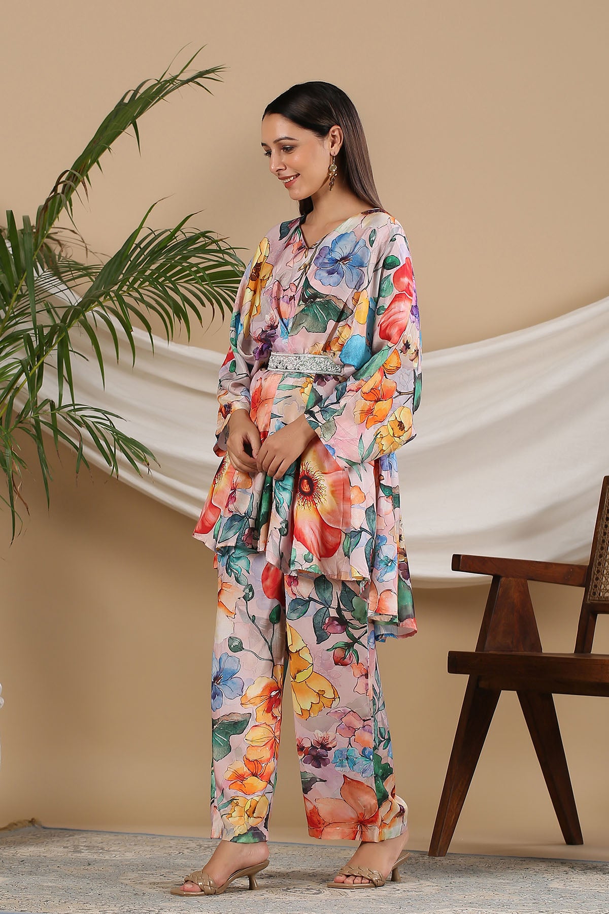 Woman wearing a colorful floral outfit standing against a beige wall with a plant and chair in the background.