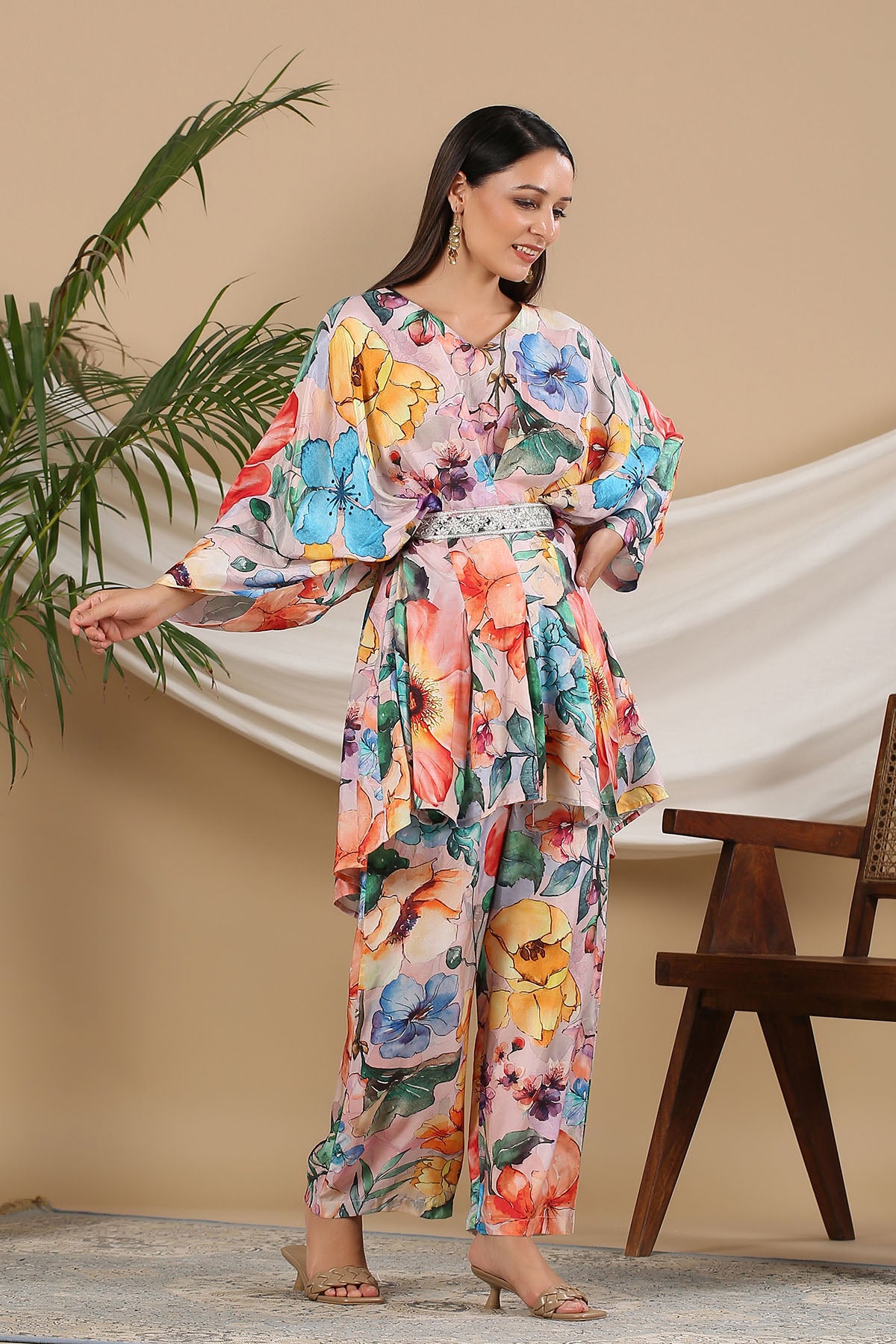 Woman wearing a colorful floral outfit standing in a room with a plant and chair.