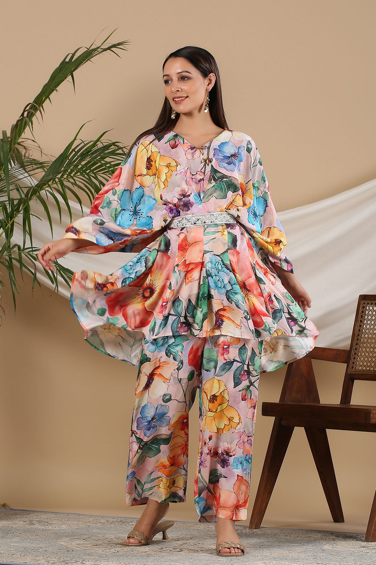 Woman wearing a colorful floral outfit standing indoors with a plant and chair in the background.