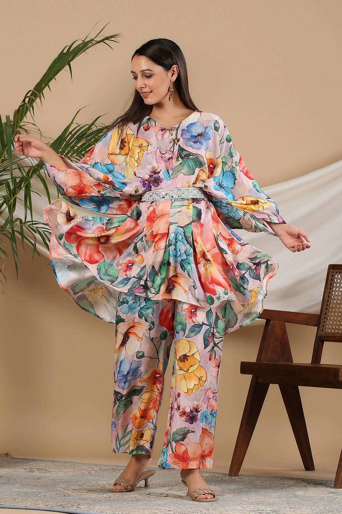 Woman wearing a colorful floral outfit standing indoors with a plant and chair in the background.