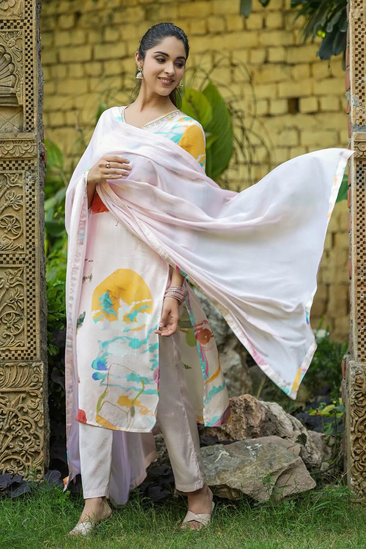 Woman in a light pink saree with colorful patterns standing outdoors.