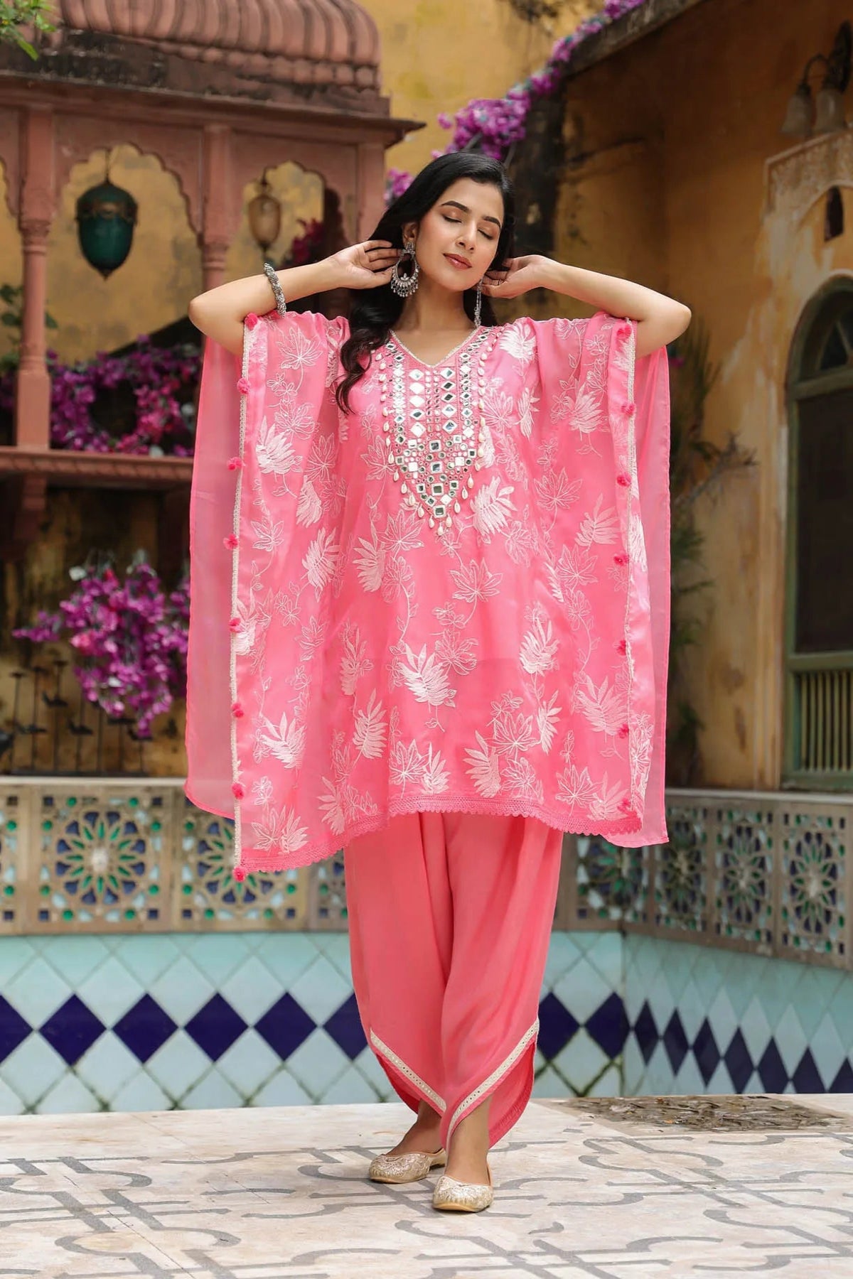Woman in a pink traditional outfit with floral patterns standing in front of a decorative wall.