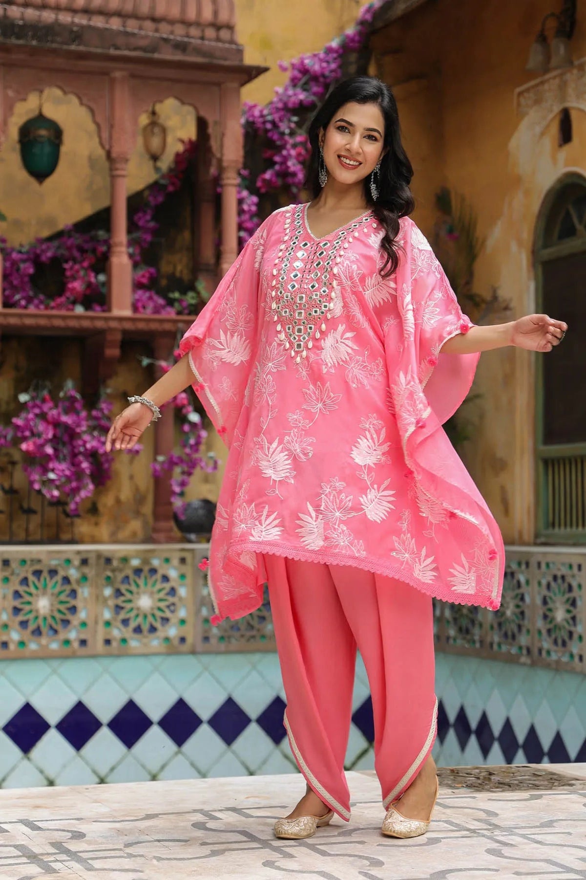 Woman in a pink traditional outfit standing in front of a decorative wall with flowers.