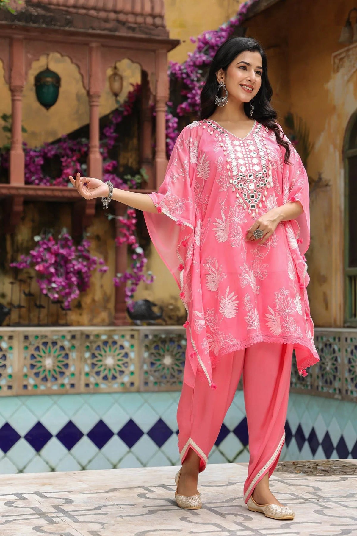 Woman in a pink traditional outfit standing in front of decorative wall and plants.