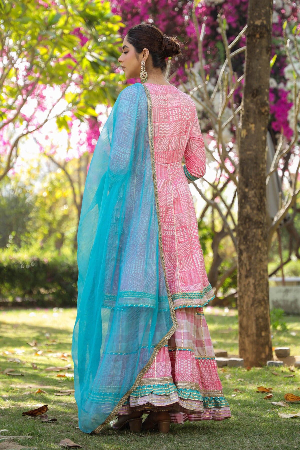 Woman in a pink traditional outfit with a blue dupatta standing in a garden with pink flowers.