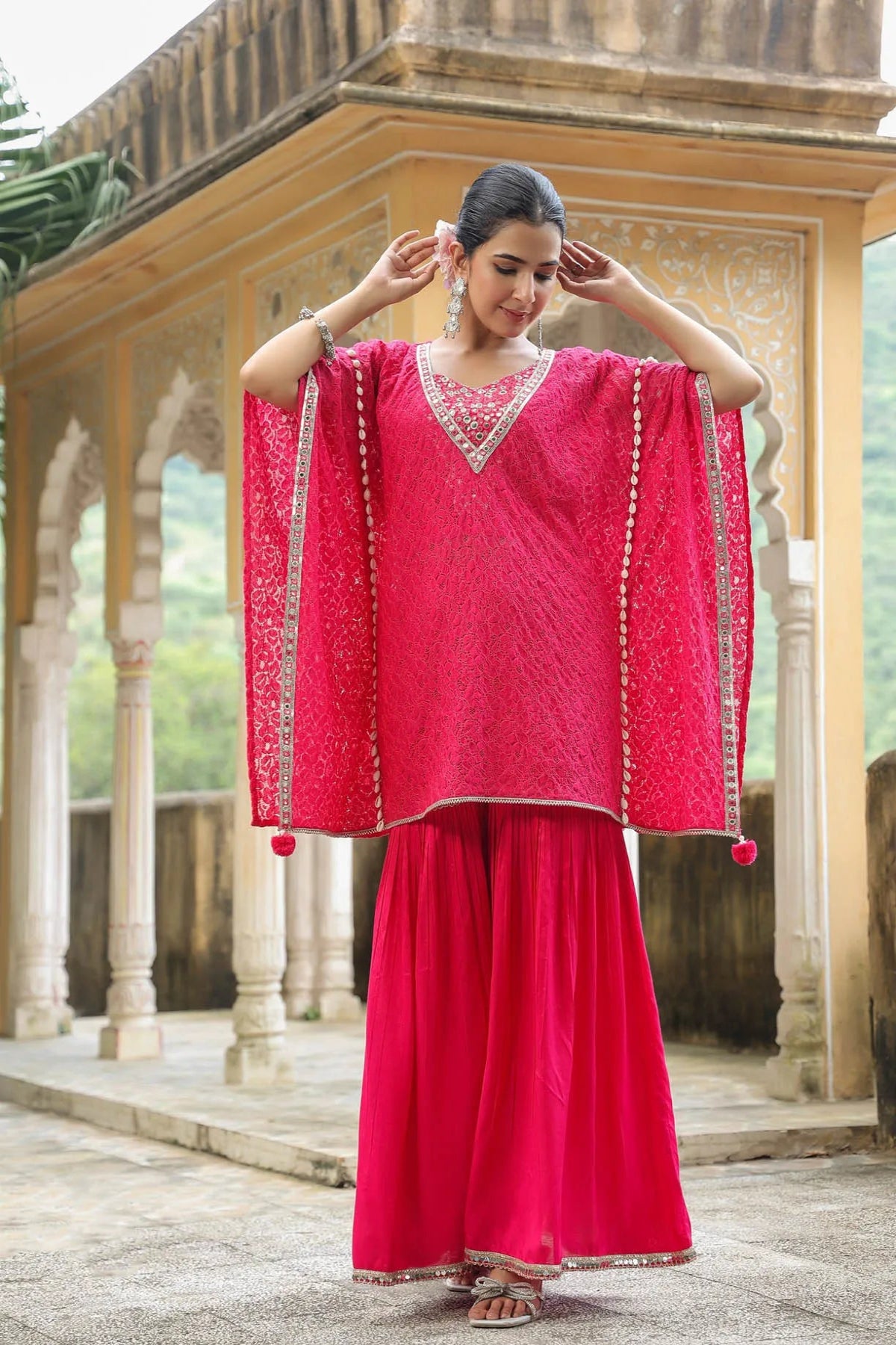 Woman in a bright pink traditional outfit with intricate patterns, standing in front of an architectural structure.