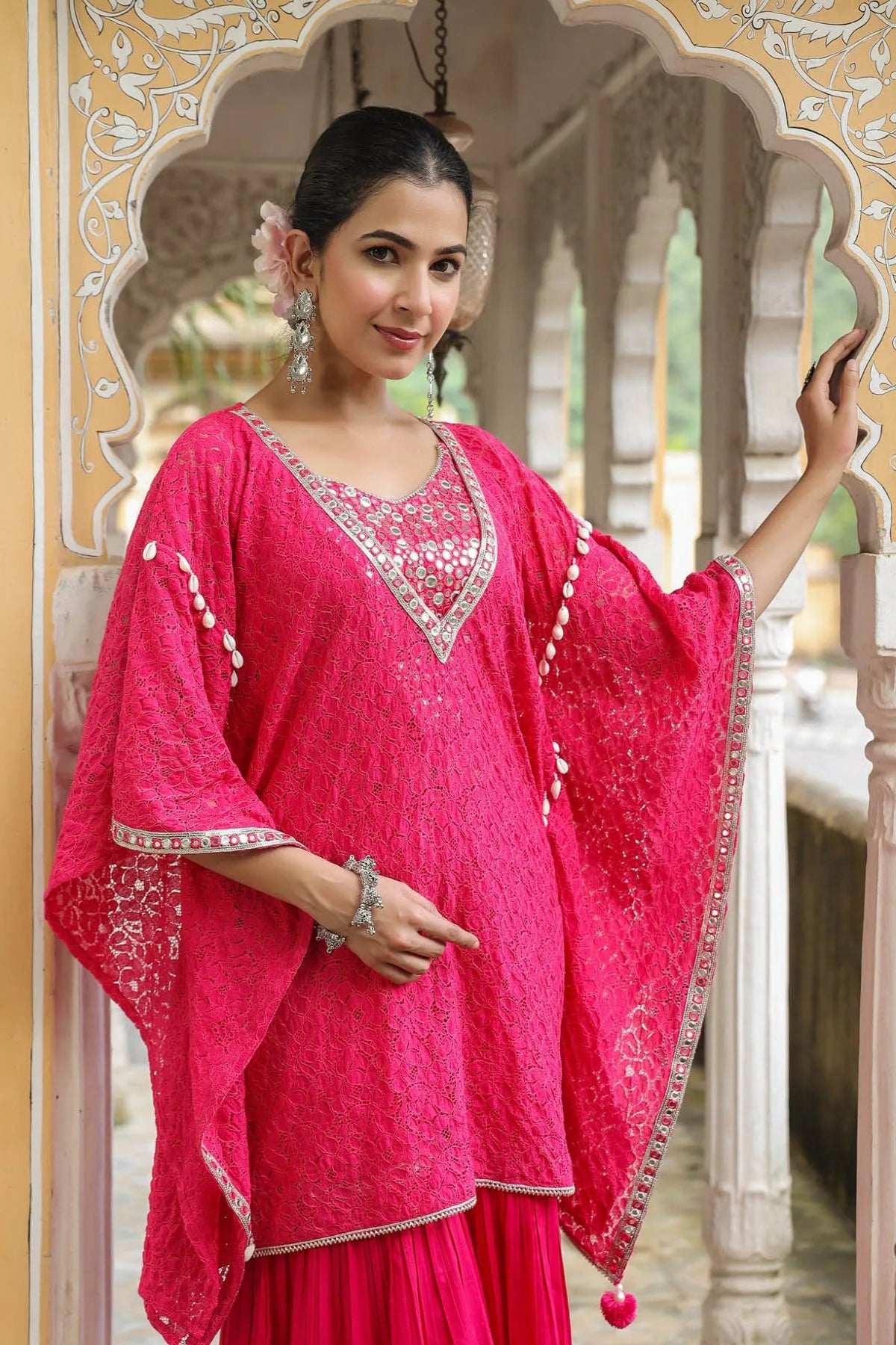 Woman in a bright pink traditional outfit with white embroidery, standing in an ornate architectural setting.