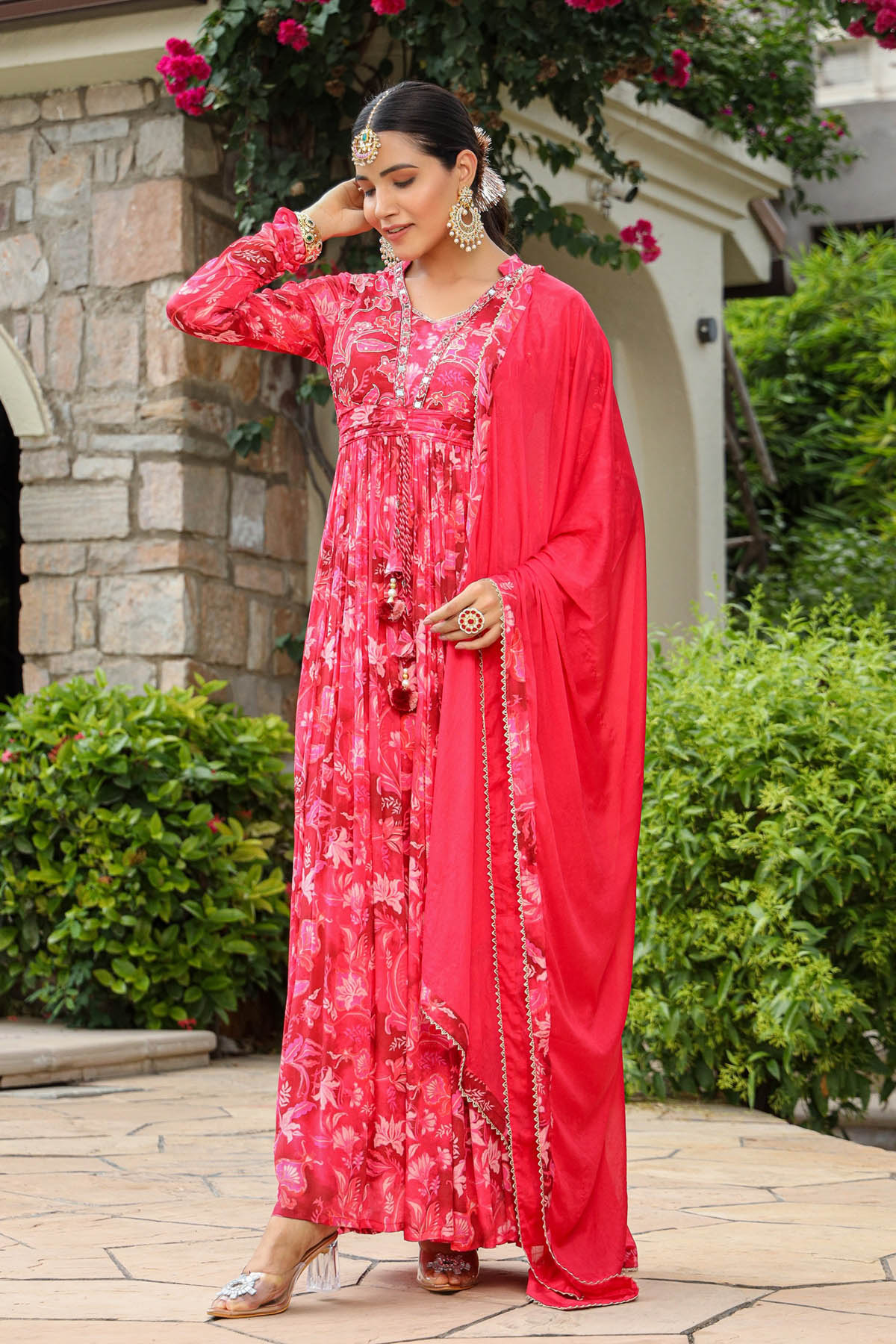 Woman in a red floral dress standing outdoors with greenery and a stone building in the background.
