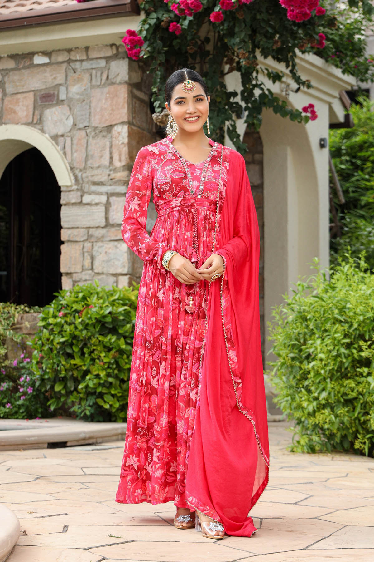 Woman in a red and pink traditional outfit standing outdoors with stone building and greenery in the background
