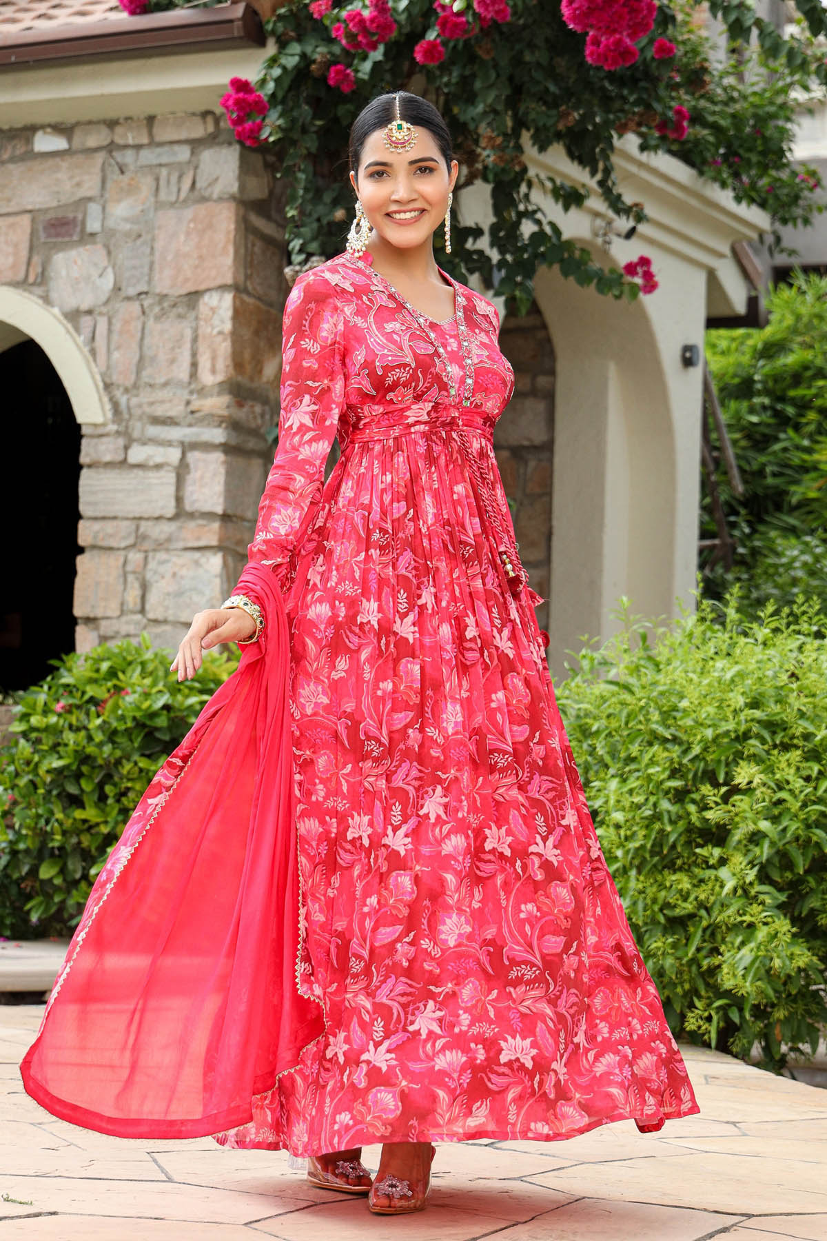 Woman in a red floral dress standing outdoors with stone building and greenery in the background