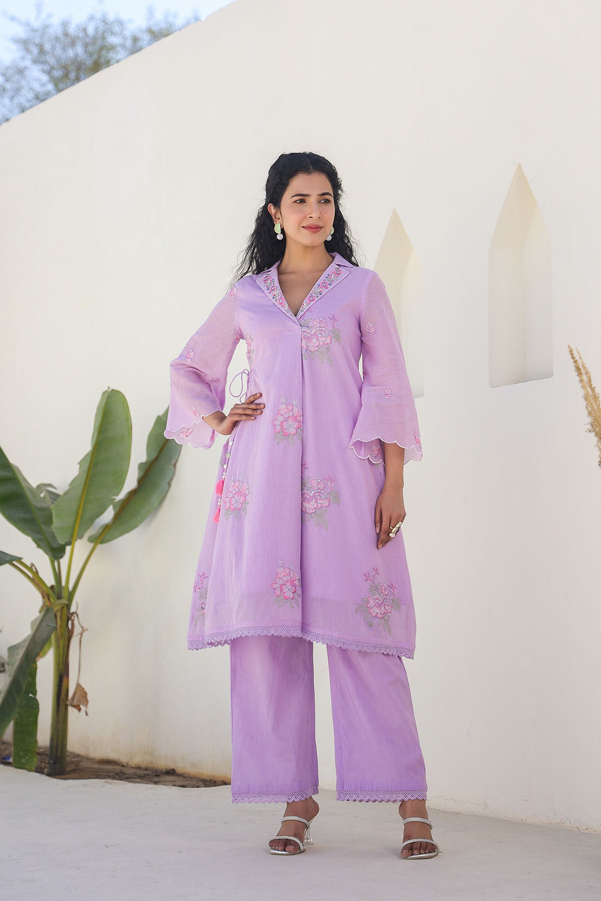 Woman wearing a light purple embroidered outfit standing against a white wall with greenery.