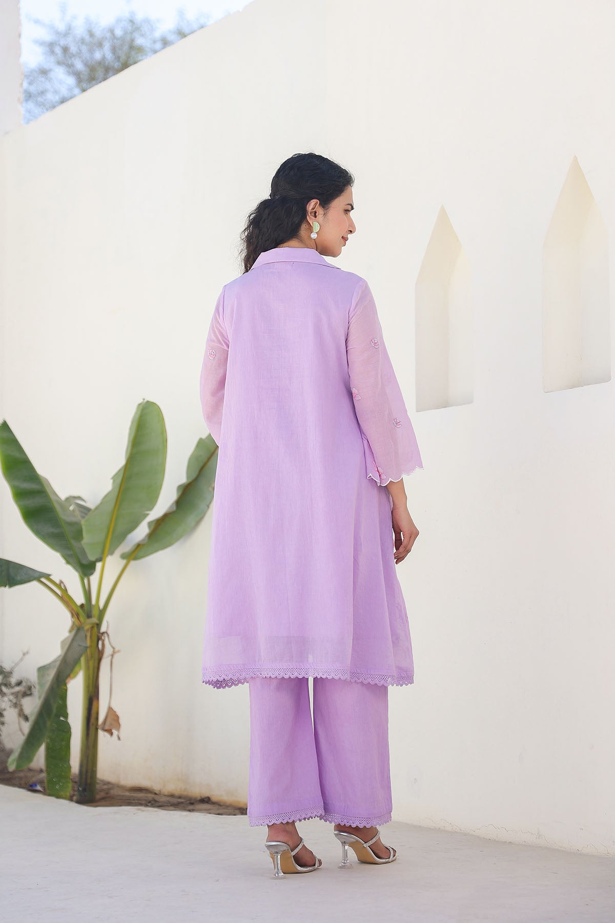 Woman wearing a light purple traditional outfit standing against a white wall with greenery.