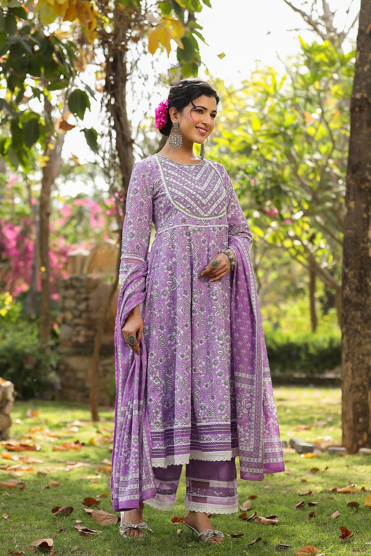 Woman in a purple traditional outfit standing in a garden with greenery and flowers.