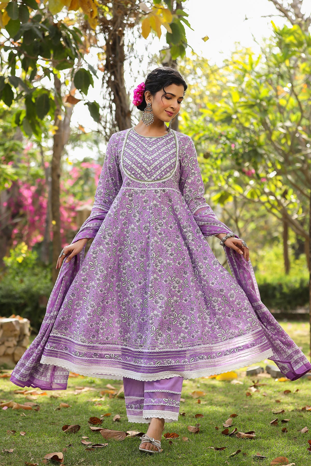 Woman in a purple traditional outfit standing in a garden with greenery and flowers.
