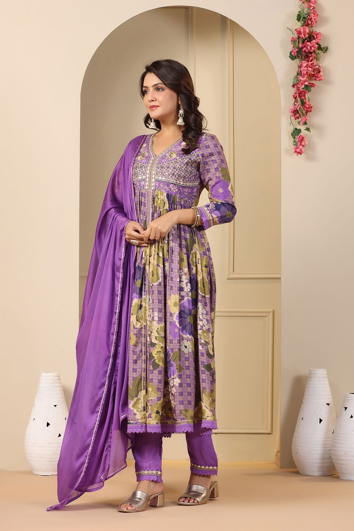 Woman wearing a purple and yellow traditional outfit with a floral pattern in a decorated room.