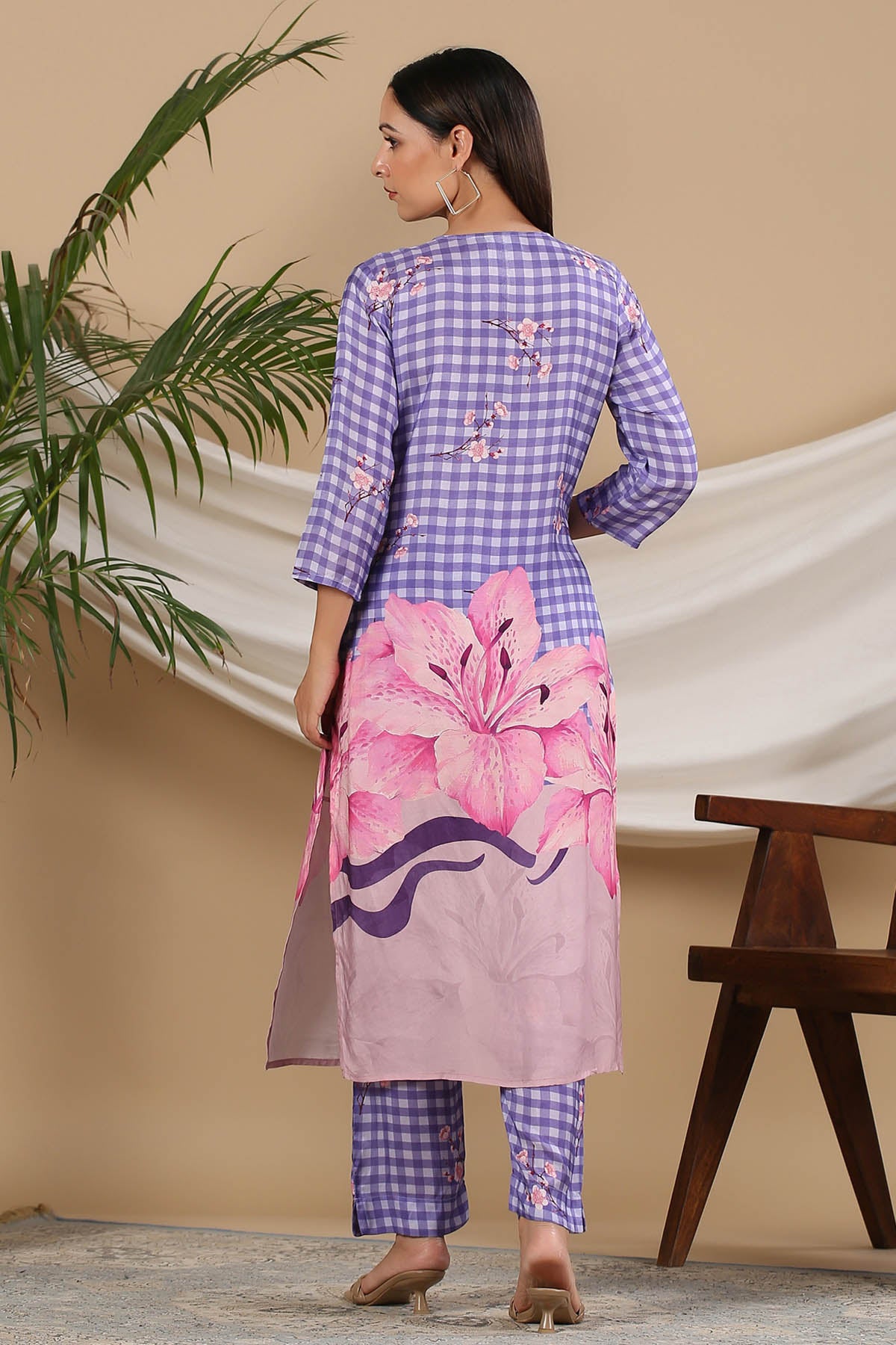 Woman wearing a purple and pink checkered kurta with floral design, standing against a beige wall.
