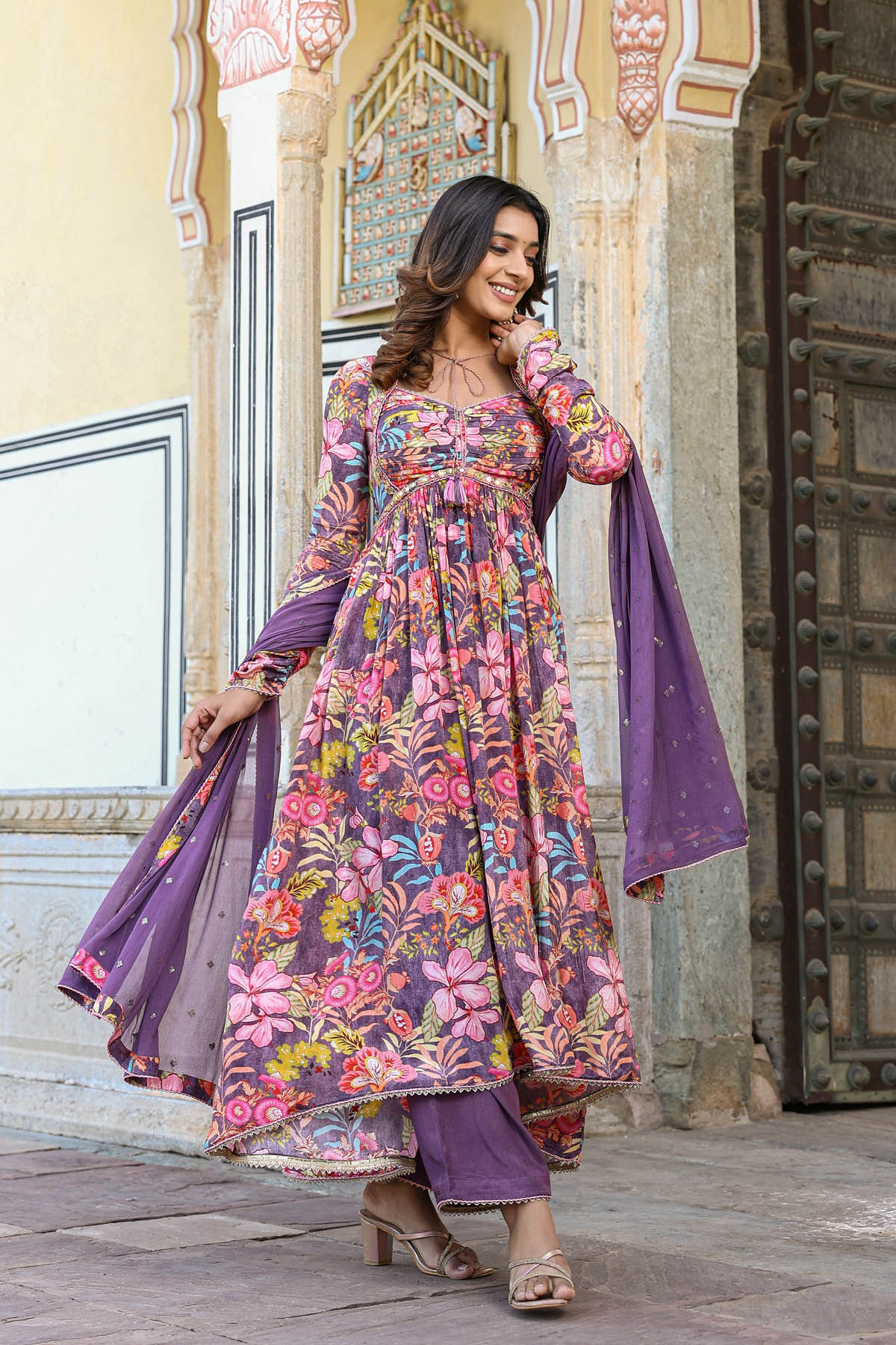 Woman in a floral dress with a purple dupatta standing in front of an ornate building.