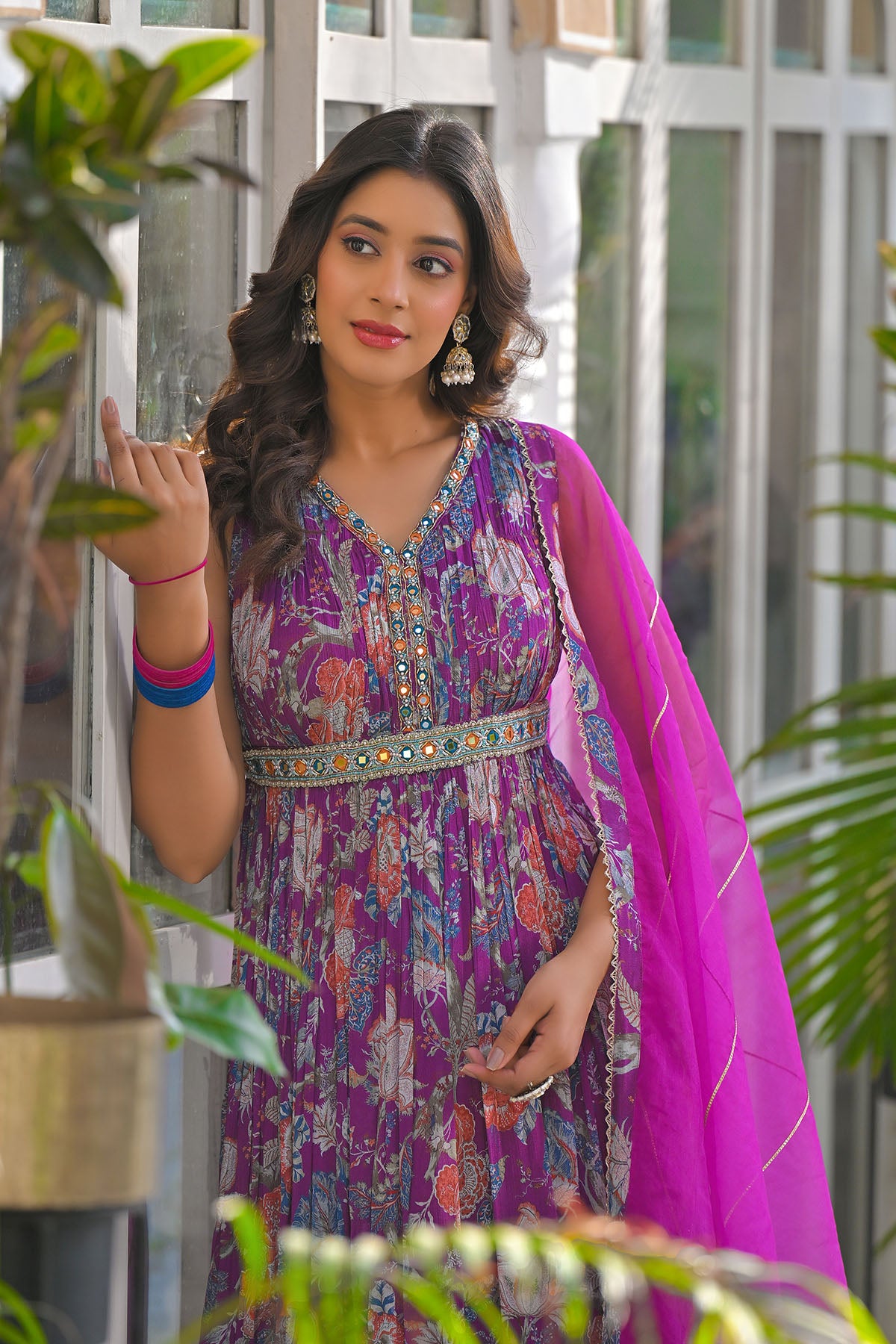 Woman in a colorful traditional outfit with a pink dupatta, standing indoors with plants around.