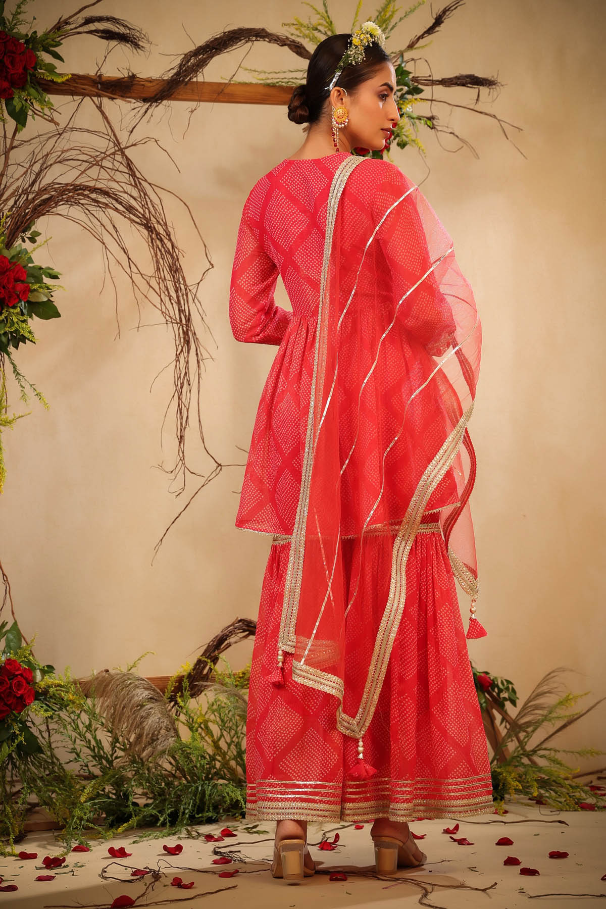 Woman in a red saree with white border standing against a decorative background with flowers and branches.