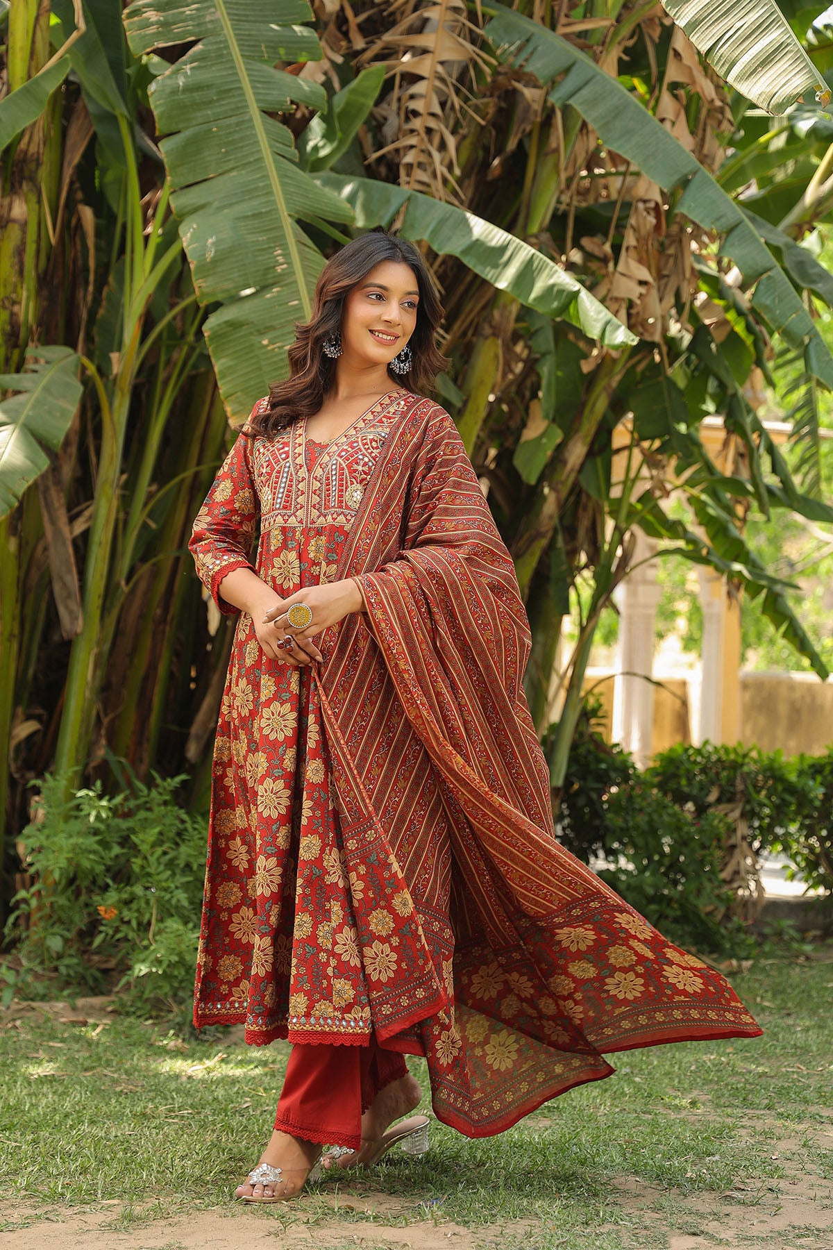 Woman in traditional red and gold outfit standing in a garden with greenery.