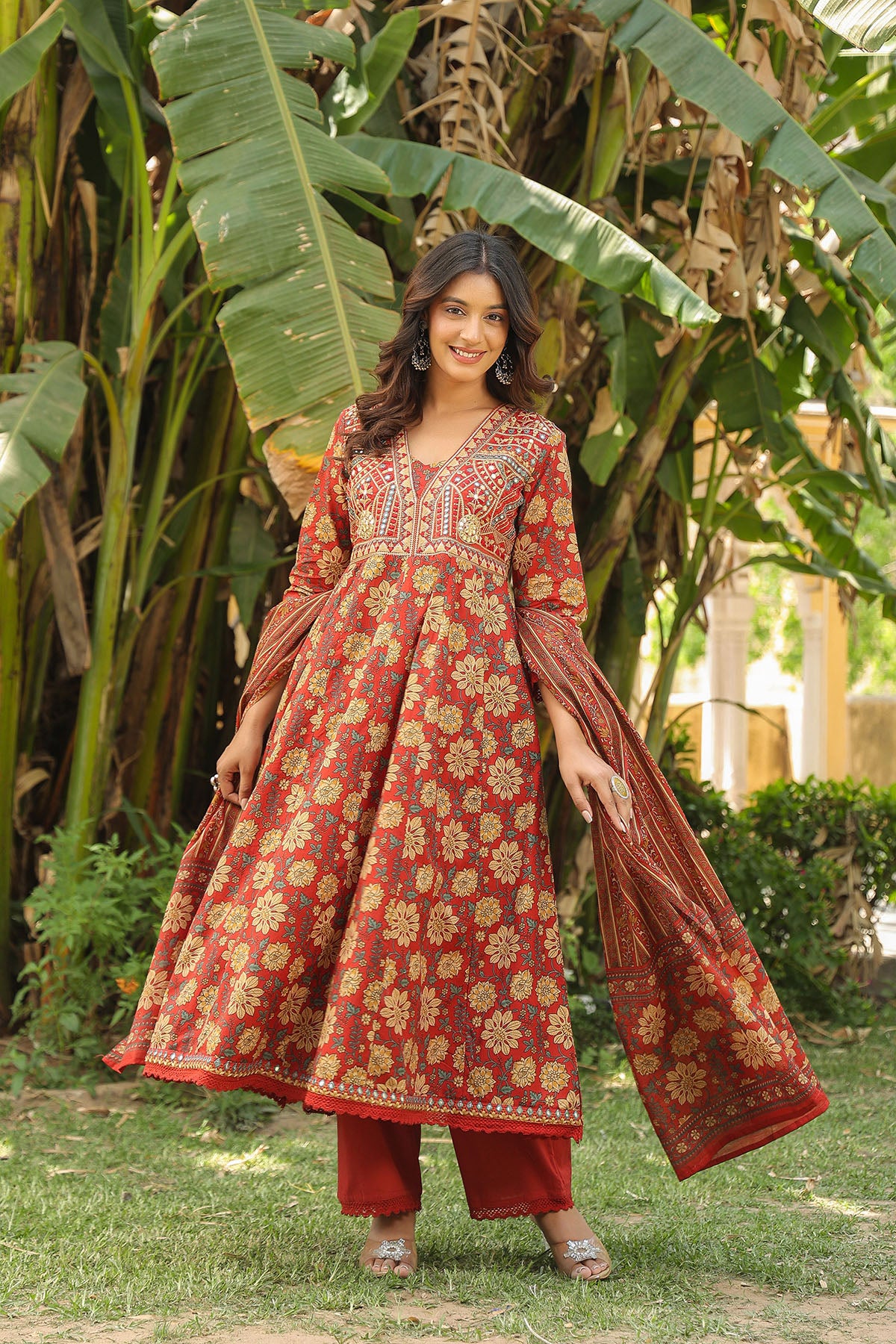 Woman in a red and gold traditional outfit standing in a garden with greenery.