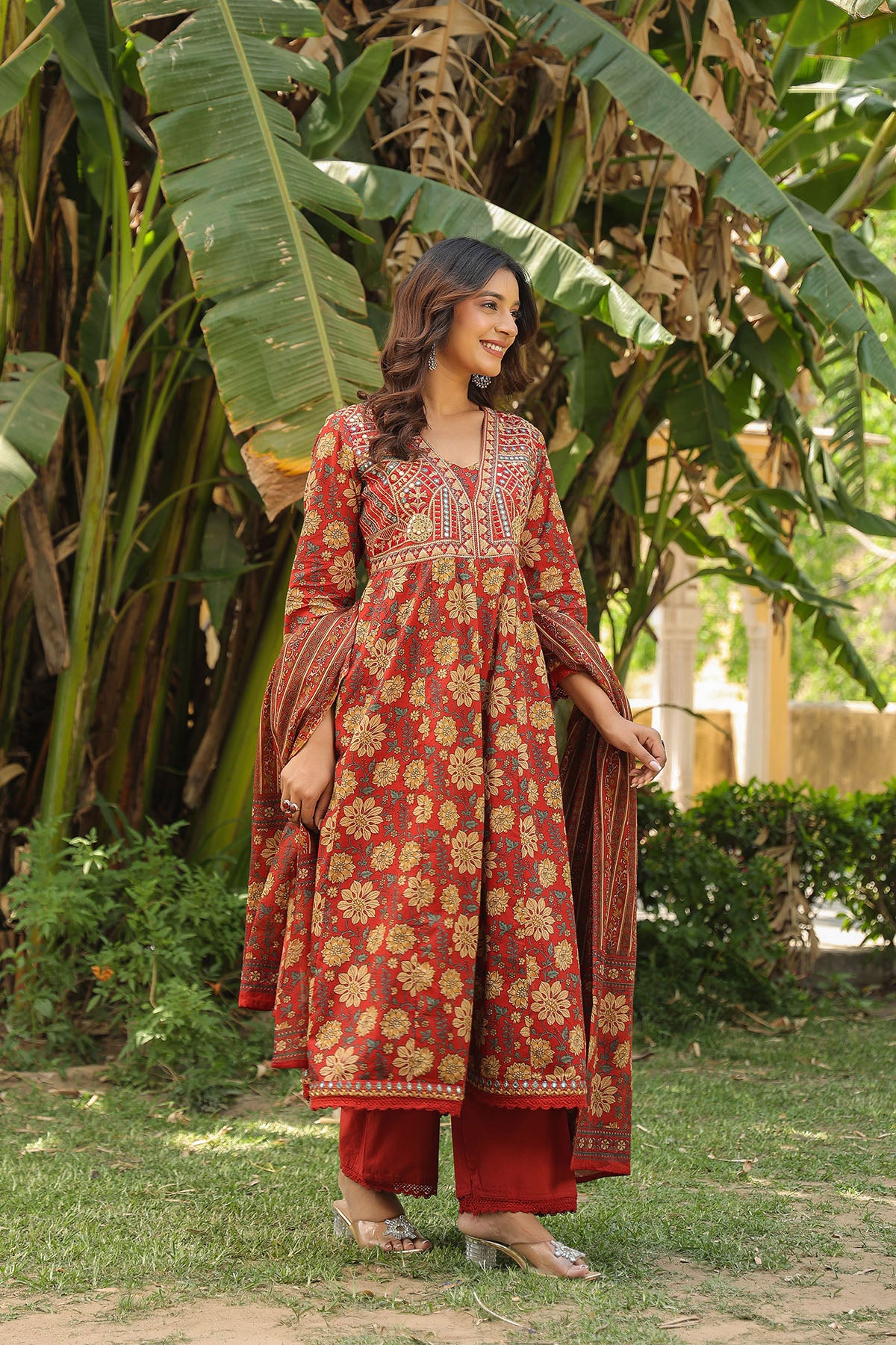 Woman in a red and gold traditional outfit standing in a garden with greenery.
