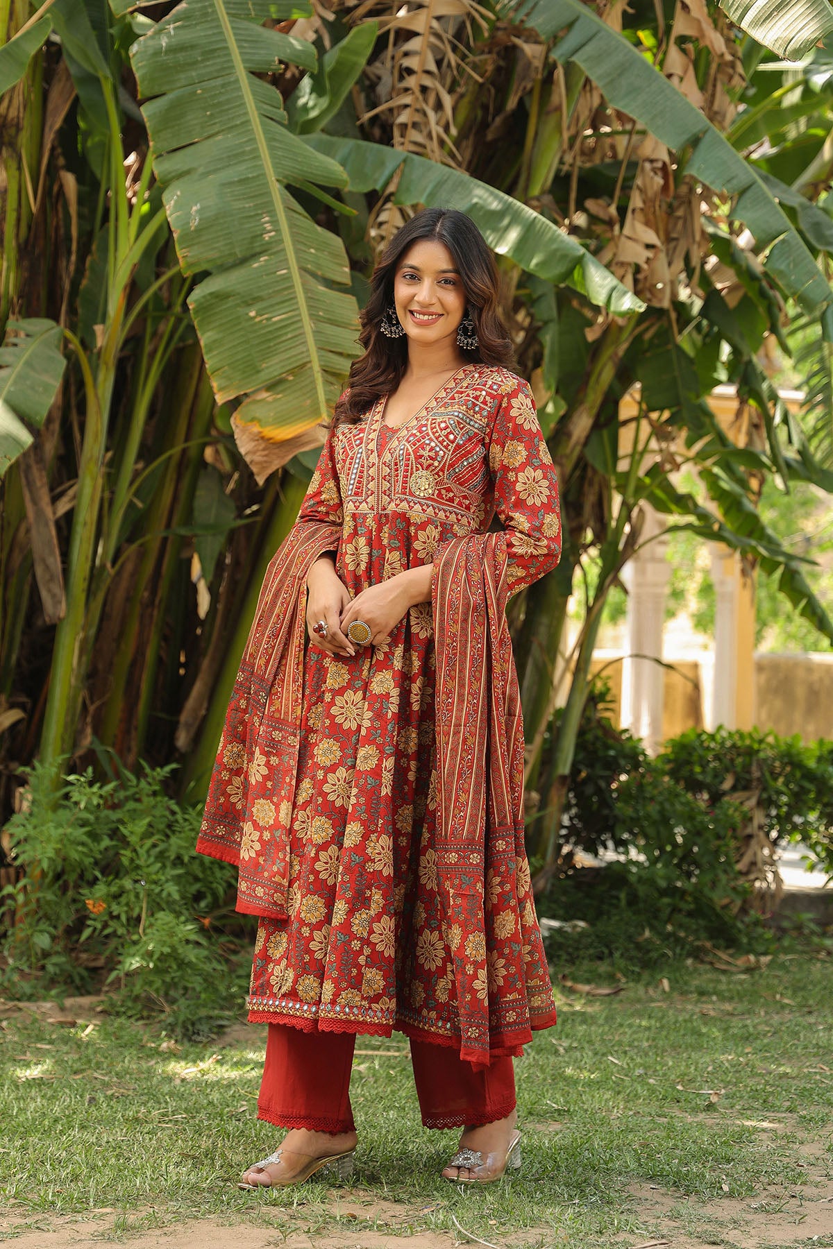 Woman in a red floral traditional outfit standing in a garden with greenery.