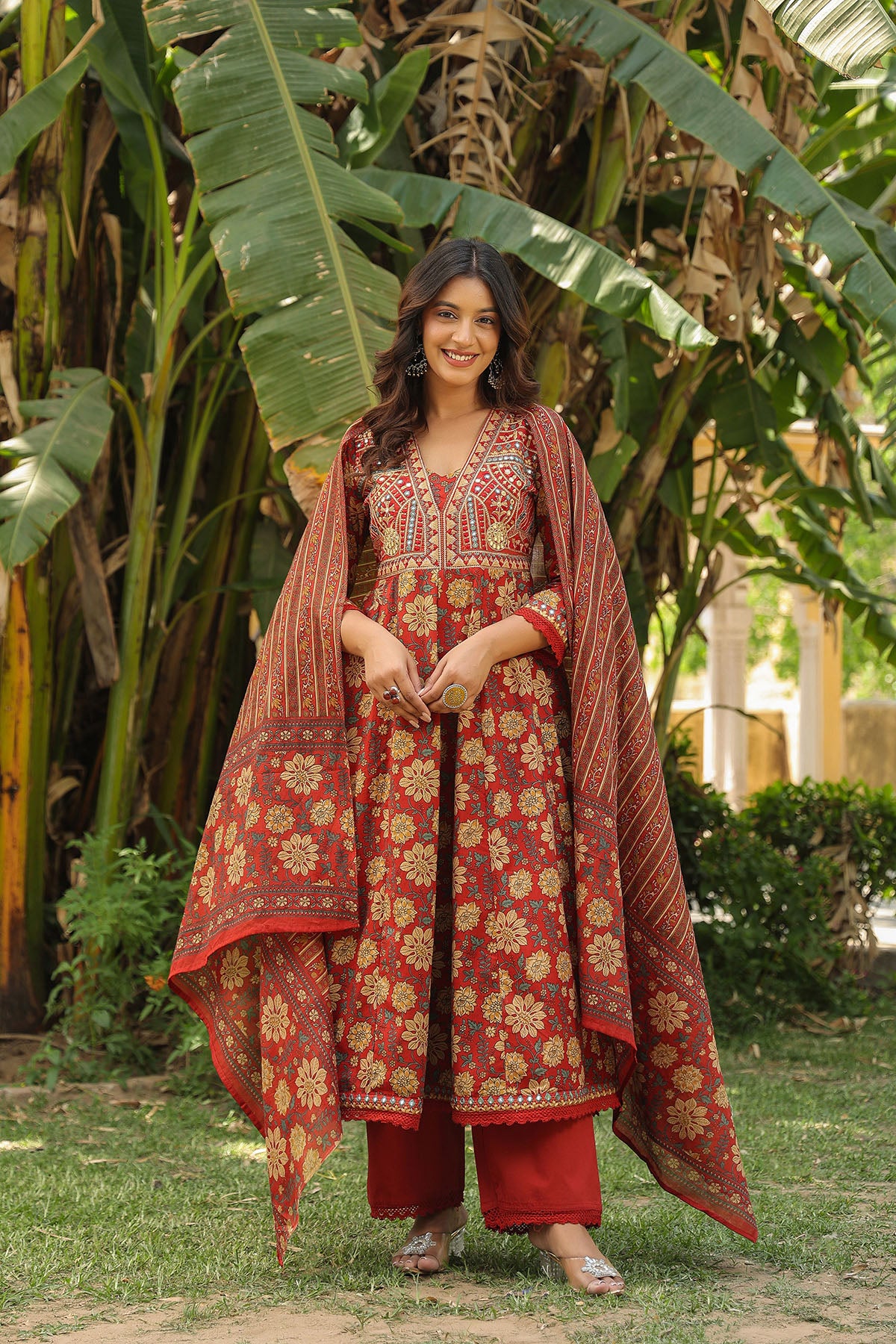 Woman in a red and gold traditional outfit standing in a garden with greenery.