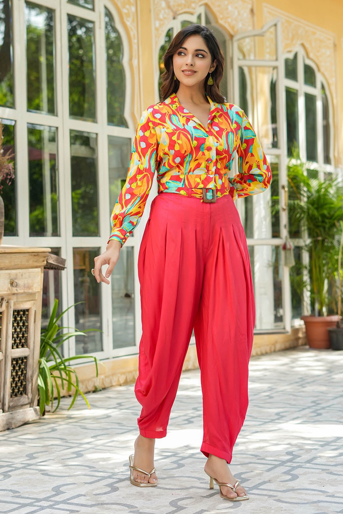 Woman wearing a colorful blouse and pink pants standing in front of a building with large windows.