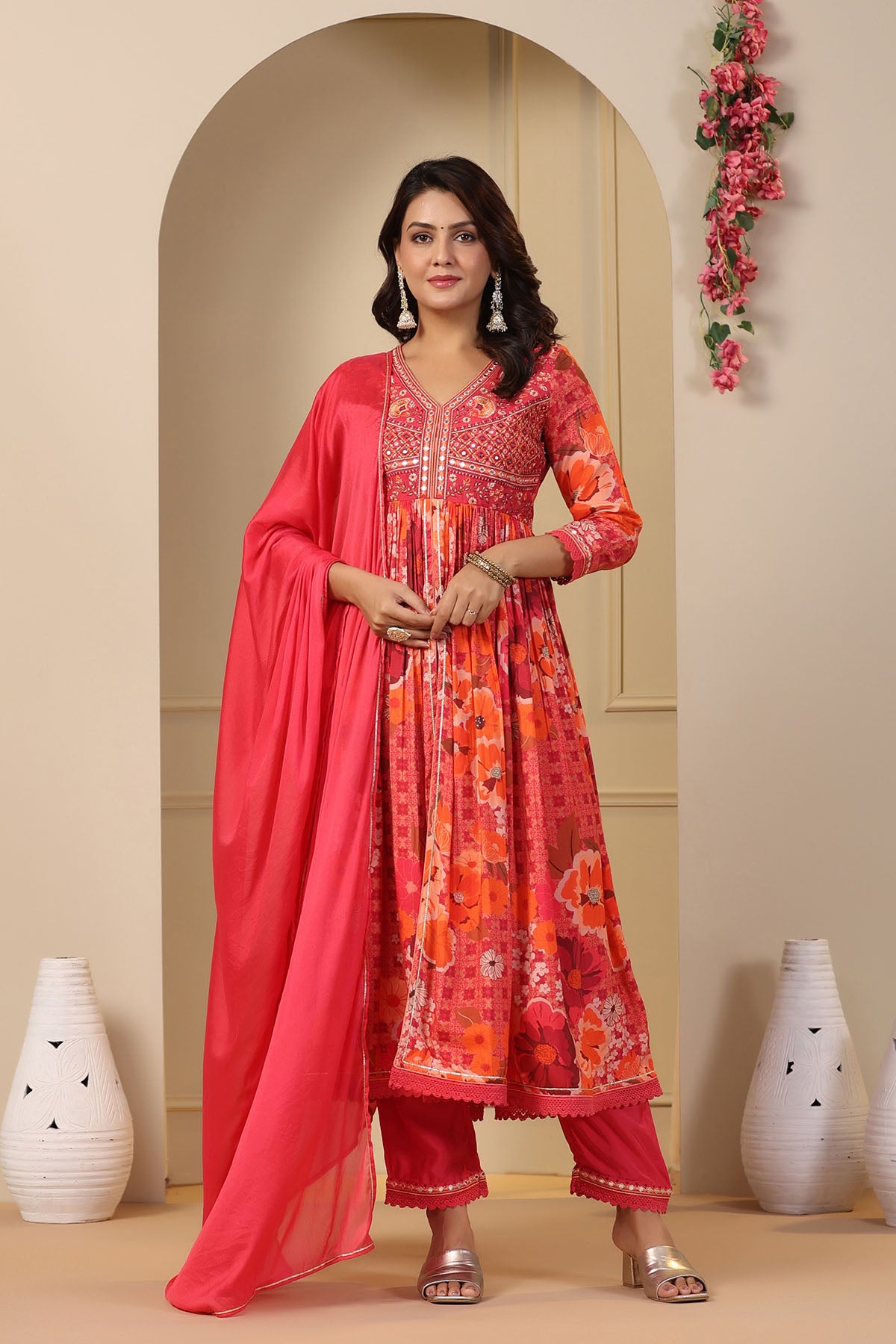 Woman in a red and orange traditional outfit with a pink dupatta standing in a decorated room.