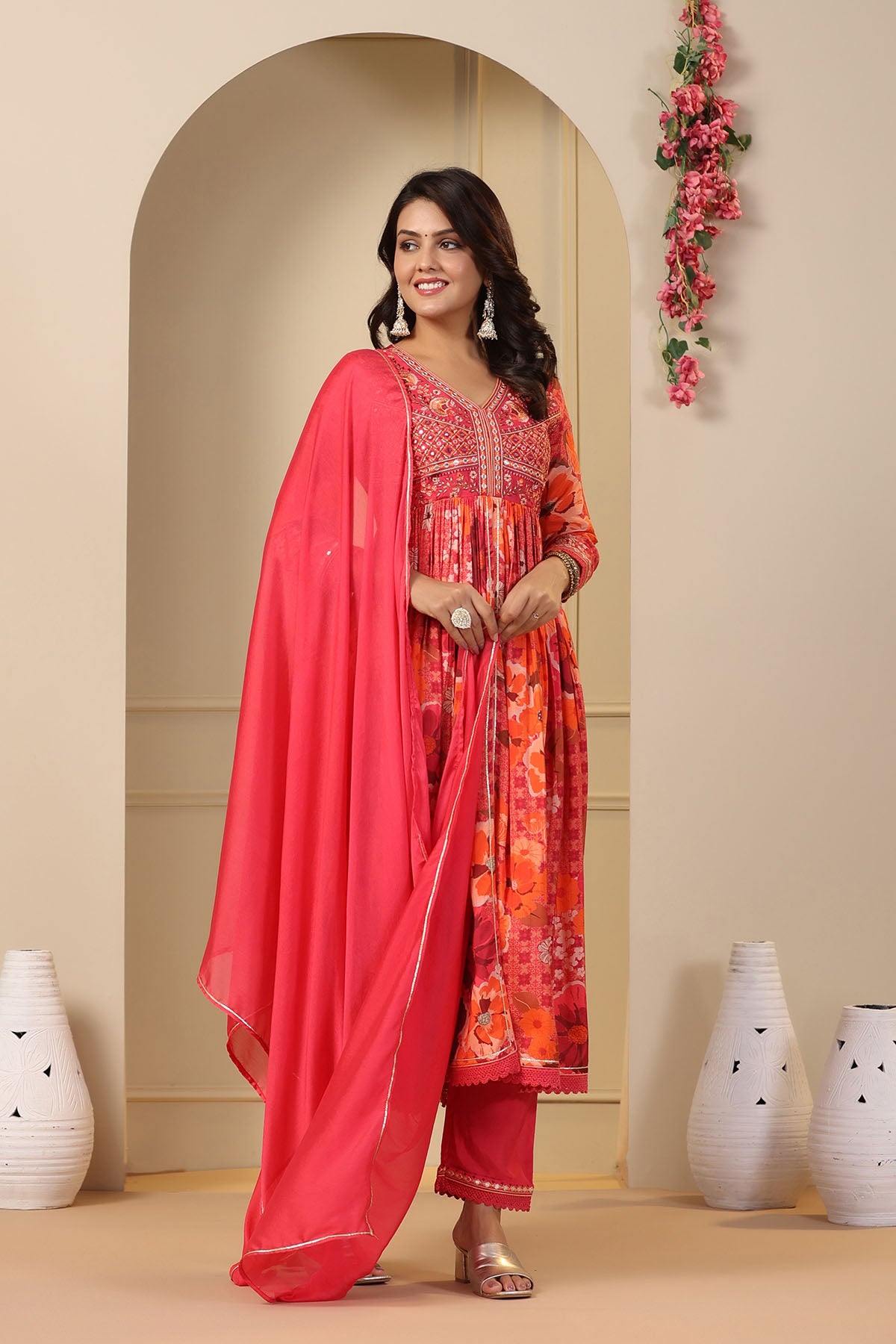 Woman in traditional outfit with pink dupatta standing in a decorated room.
