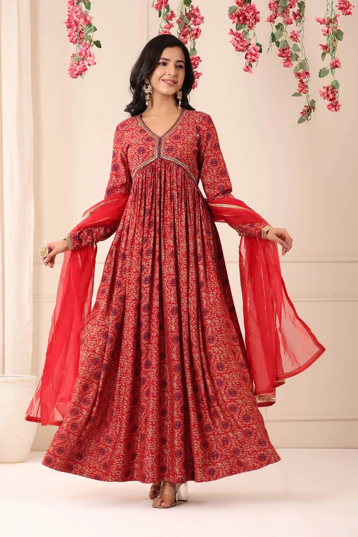 Woman wearing a red patterned dress with a sheer dupatta against a floral decorated wall.