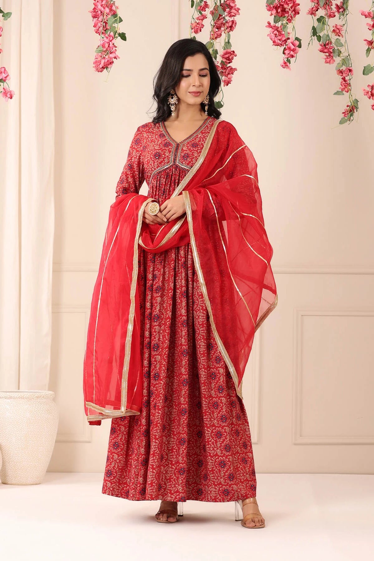 Woman in a red traditional outfit with a matching dupatta against a floral decorated wall.