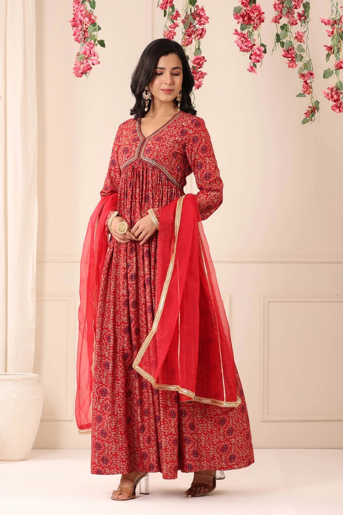 Woman wearing a red traditional outfit with a matching dupatta against a floral decorated wall.