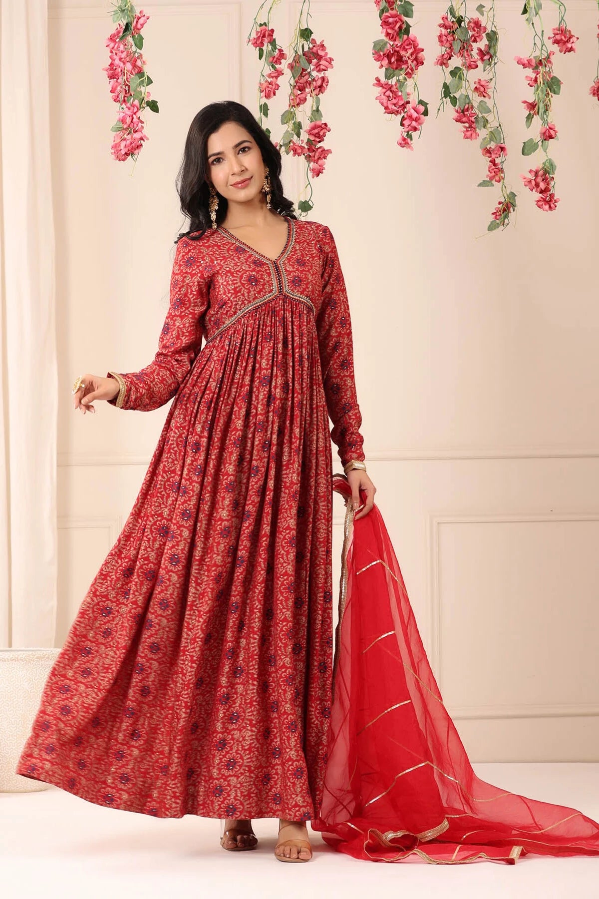 Woman in a red patterned dress with a matching dupatta against a floral decorated wall.