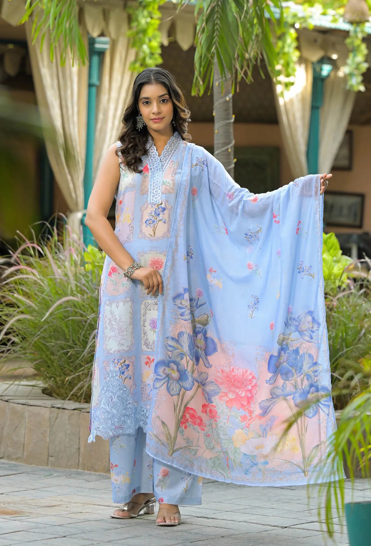 Woman holding a floral-patterned light blue dupatta in an outdoor setting with plants and curtains.