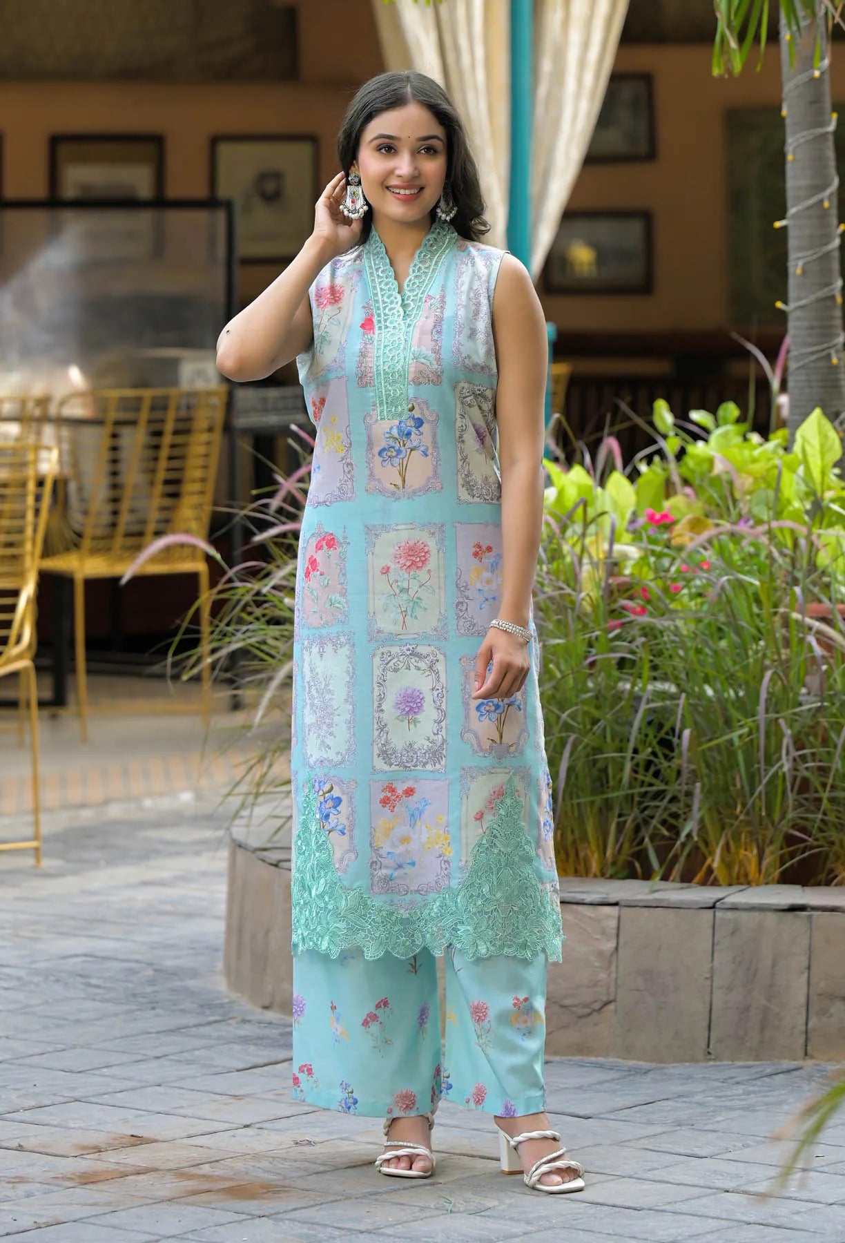 Woman in a light blue embroidered outfit standing outdoors with plants and furniture in the background
