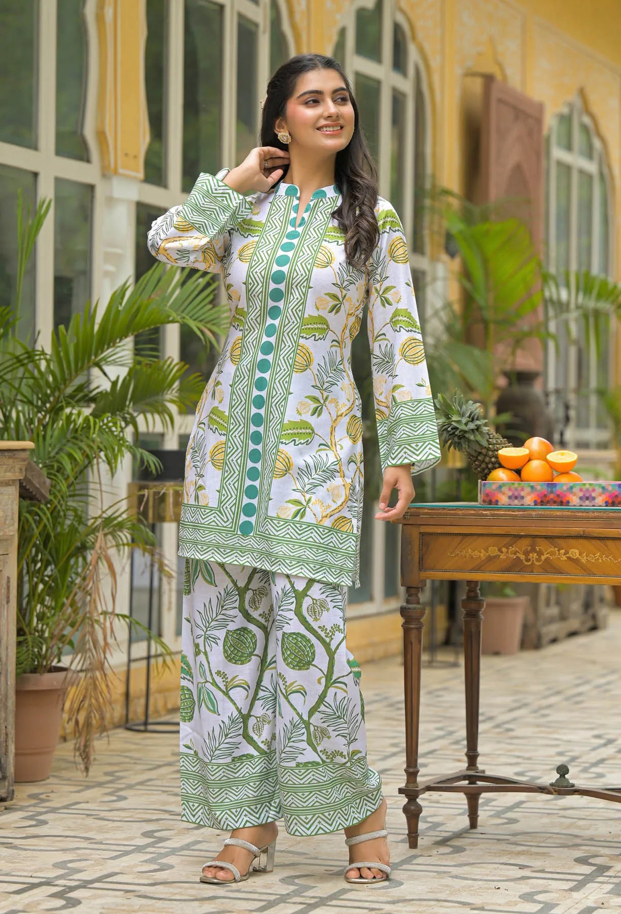 Woman in a green and white patterned outfit standing outdoors with plants and a table in the background.